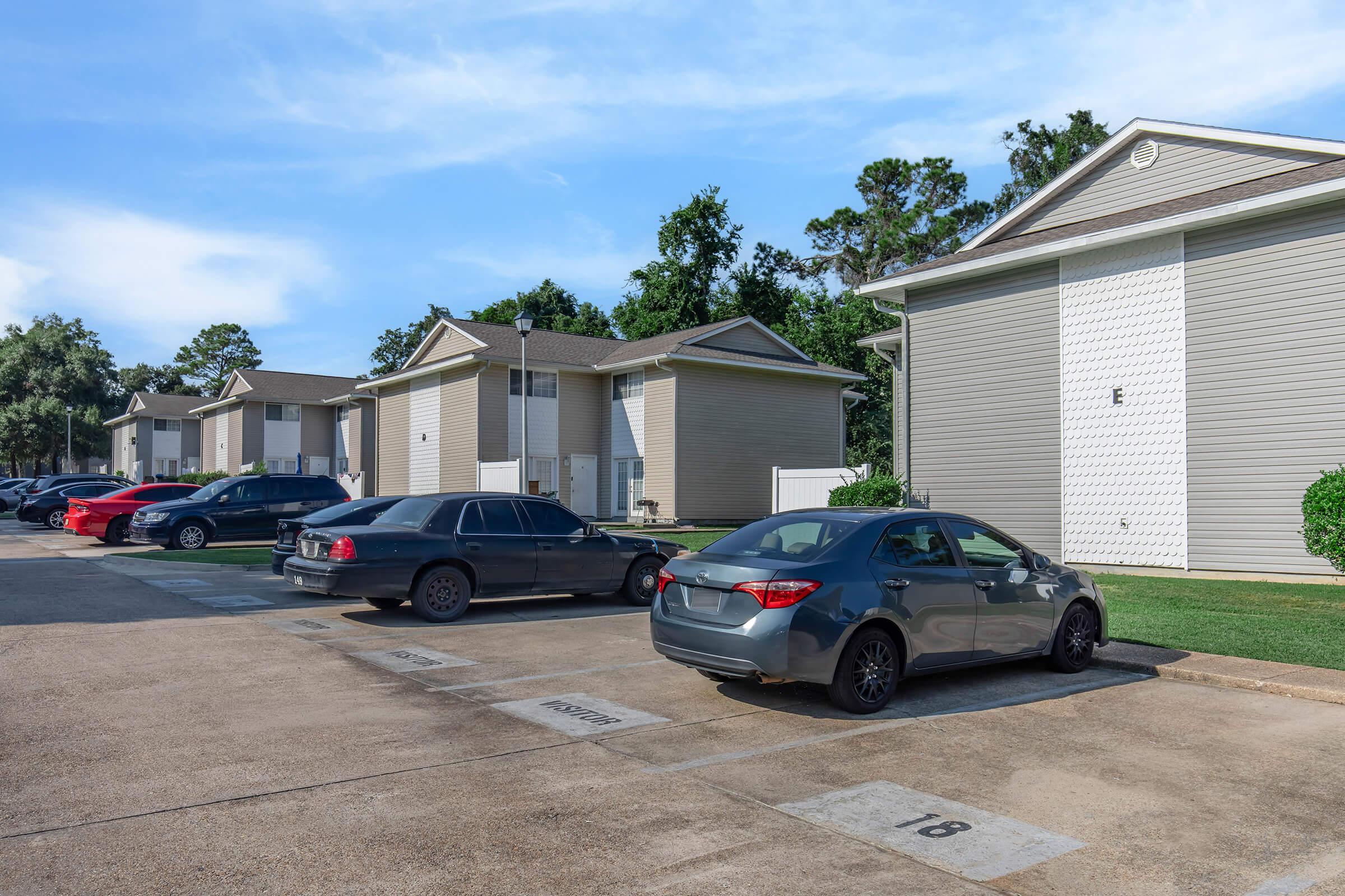 A parking lot with several vehicles, including a gray sedan and a black car, adjacent to residential buildings. The scene features green grass, trees in the background, and clear blue skies. Parking spots are marked with numbers, indicating designated areas for cars.