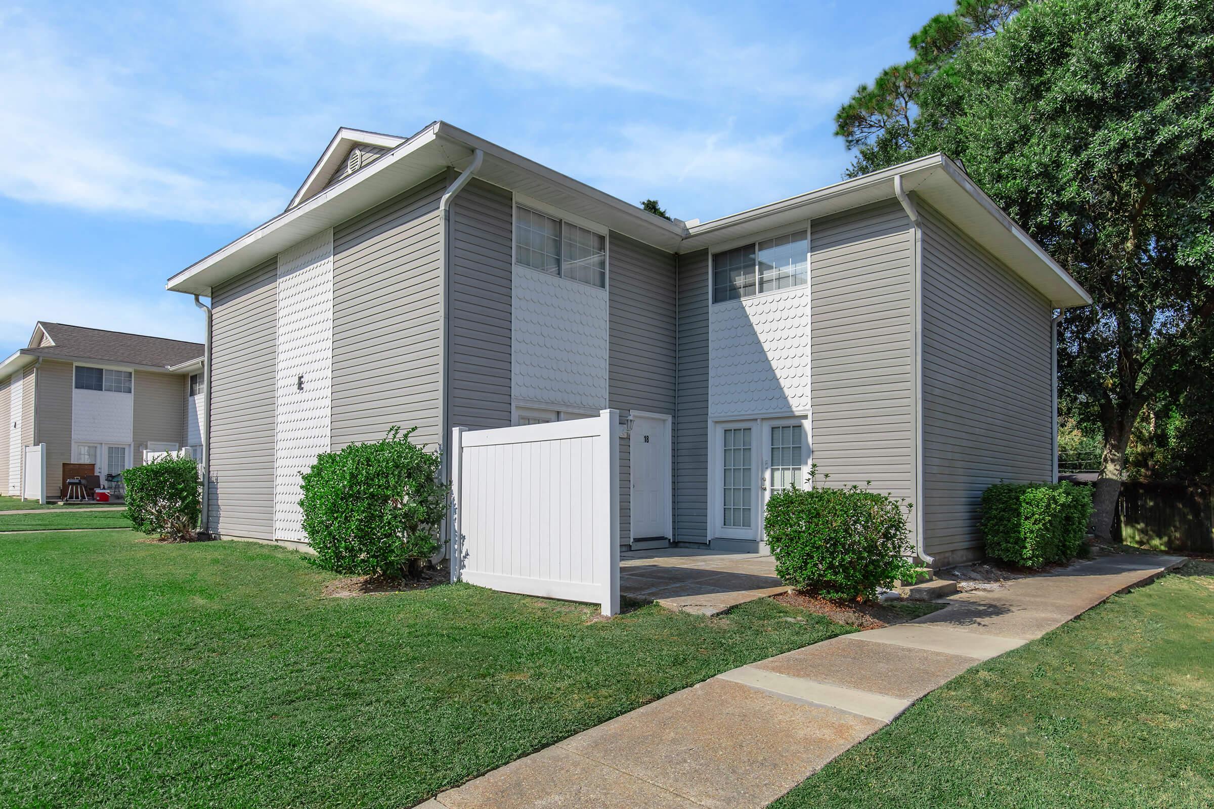 A two-story residential building with light gray siding and white trim, featuring multiple windows. The entrance is framed by green shrubs and a white fence, with a sidewalk leading to the door. Lush green grass surrounds the property, and the sky is clear and blue in the background.