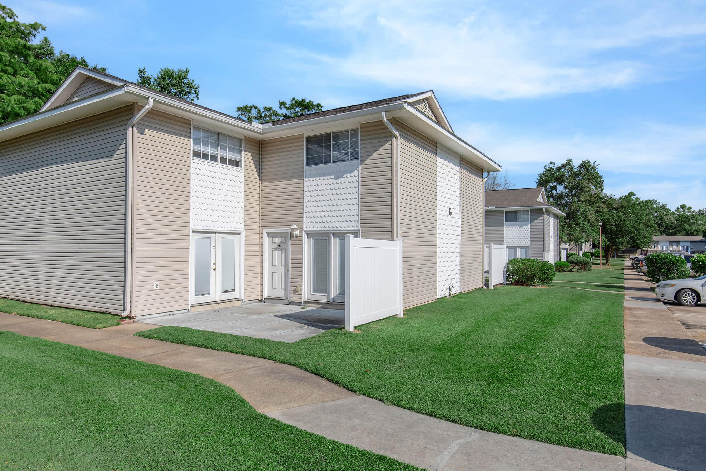 Exterior view of a two-story residential building featuring light-colored siding. The building has a small porch area and large windows. Well-maintained green grass surrounds the structure, with a walking path and several nearby similar buildings visible in the background under a clear blue sky.