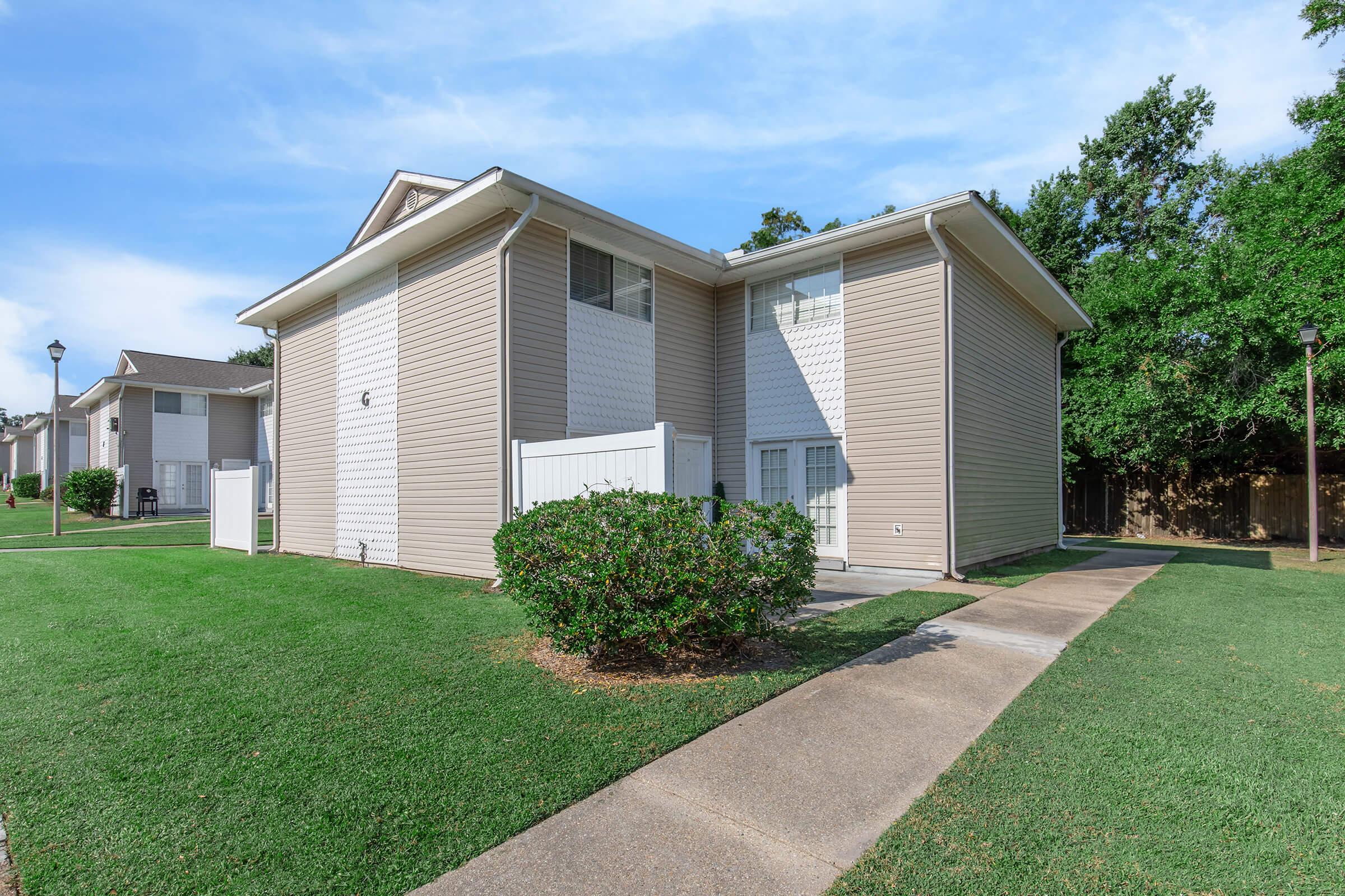 A two-story residential building with beige siding, featuring a well-manicured lawn and shrubs. The pathway leads to the entrance, with a white fence on the side. Trees are visible in the background, under a clear blue sky.