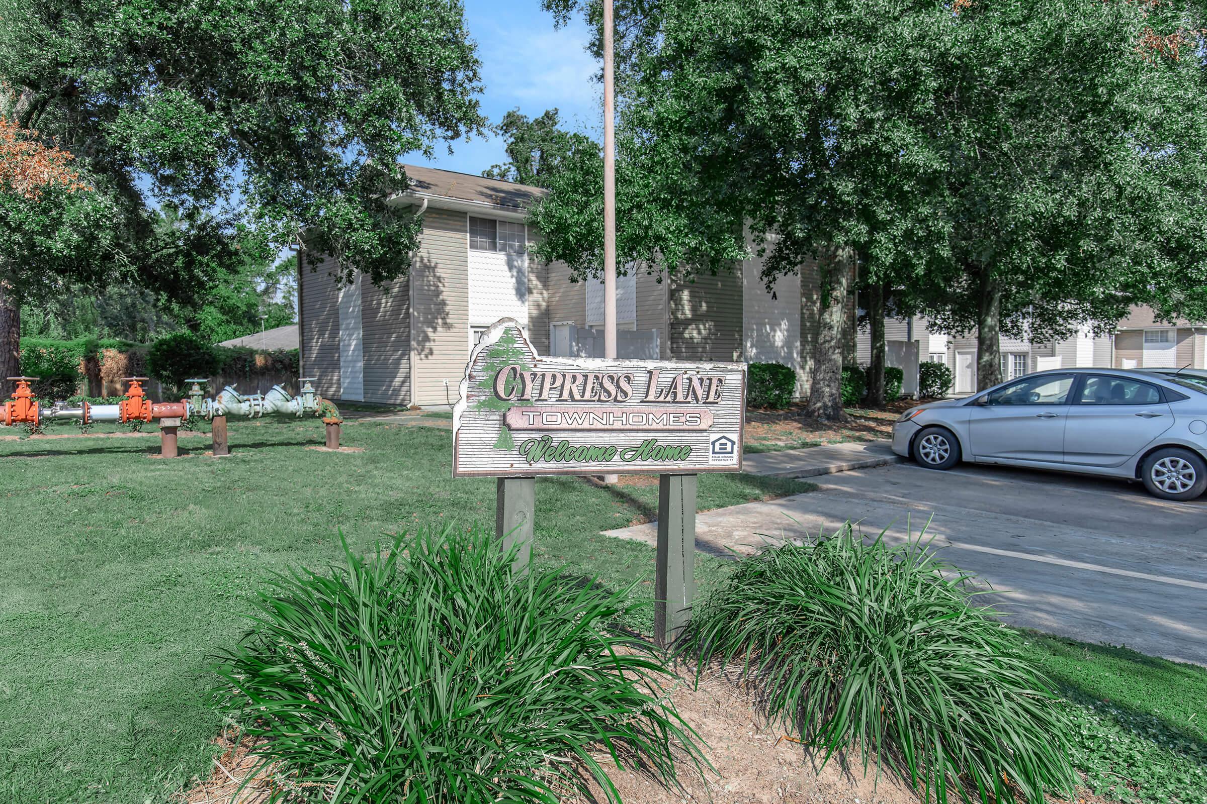 Sign for Cypress Lane Townhomes, featuring the community name prominently displayed. Surrounding greenery includes bushes and trees. In the background, there are residential buildings and a parked car visible on the driveway. The scene is well-lit, indicating a sunny day.