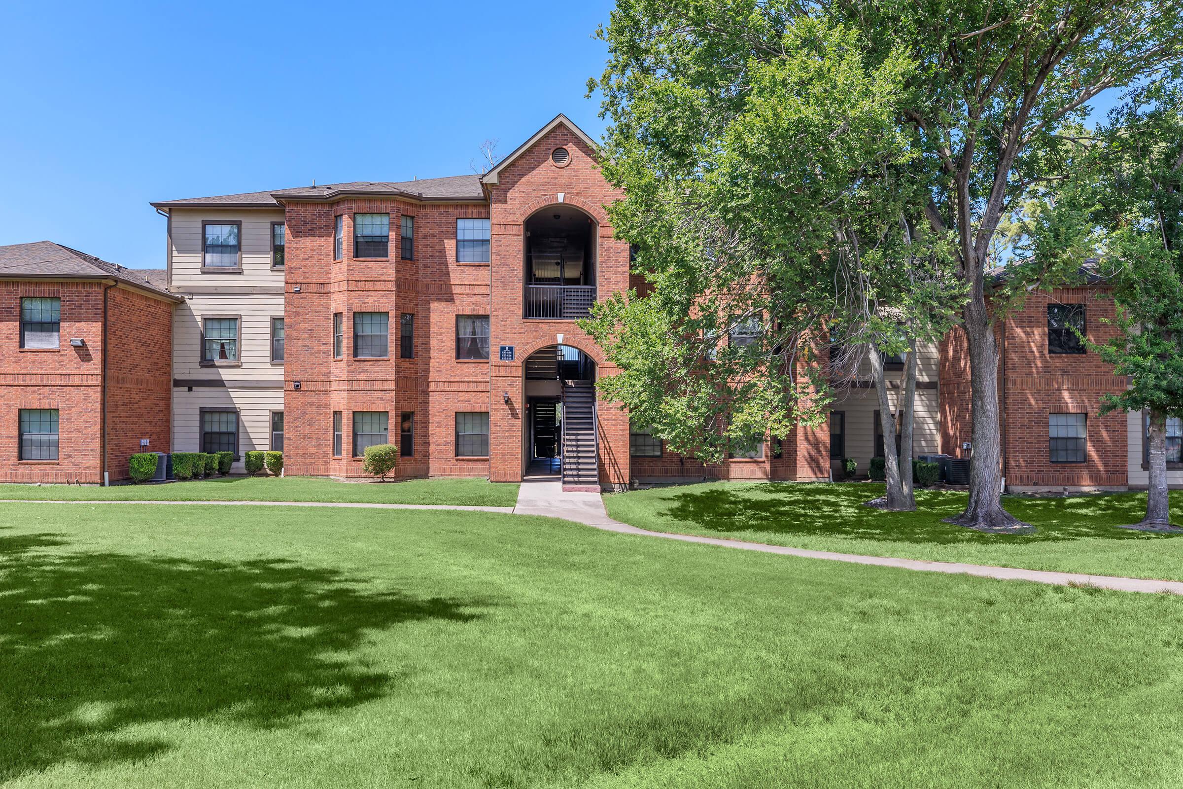 A three-story brick apartment building surrounded by greenery. The building features multiple balconies and large windows, with a winding path leading to the entrance. Trees and manicured lawns enhance the inviting atmosphere of the property on a clear, sunny day.