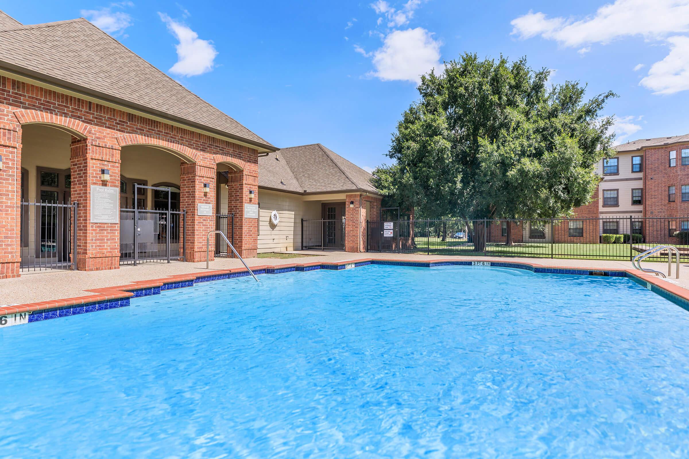 A clear blue swimming pool with a shallow end marked at 3 feet and a deep end at 6 feet. Surrounding the pool are brick buildings and lush green trees. The area is sunny with a light blue sky, creating a welcoming outdoor space.