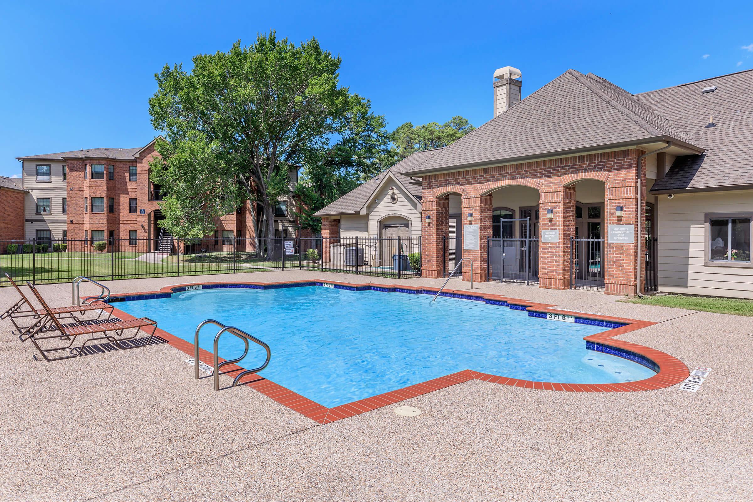 A clear blue swimming pool surrounded by a patio area with lounge chairs. In the background, there are residential buildings and lush green trees under a bright blue sky. The pool area has a safety fence and includes steps for entry.