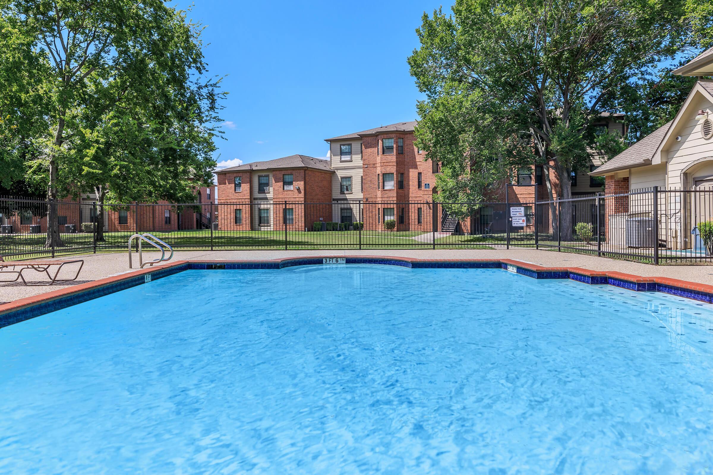 A clear blue swimming pool surrounded by green trees and a lawn, with brick apartment buildings in the background. The scene is sunny, showcasing a relaxing outdoor space ideal for leisure and recreation.