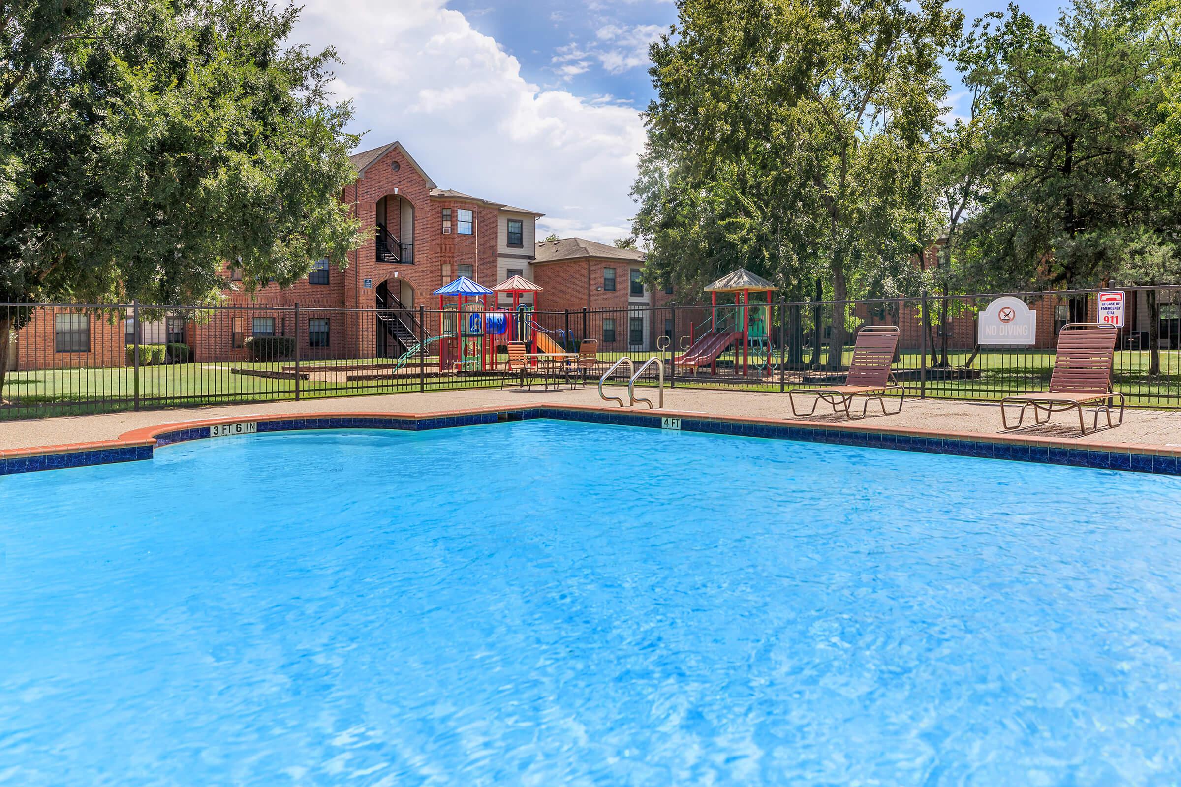 A bright blue swimming pool with lounge chairs sits in the foreground. In the background, there are brick apartment buildings and a playground with slides and swings. Lush green trees surround the area, creating a relaxing outdoor space. The sky is partly cloudy, adding to the serene atmosphere.
