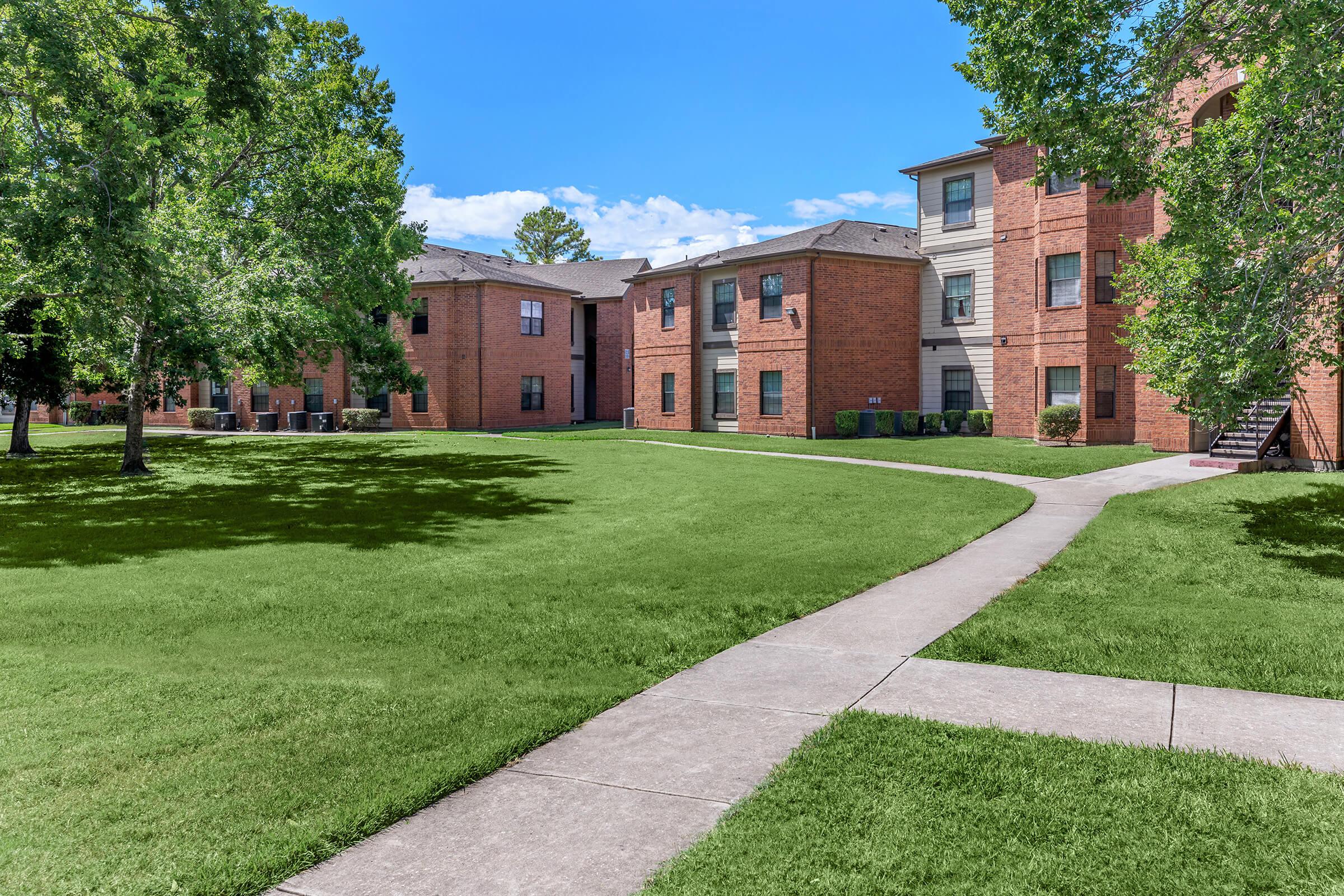 A well-maintained residential landscape featuring several brick apartment buildings surrounded by green lawns and shade trees. A concrete pathway winds through the grass, leading to the entrances of the apartments under a clear blue sky.