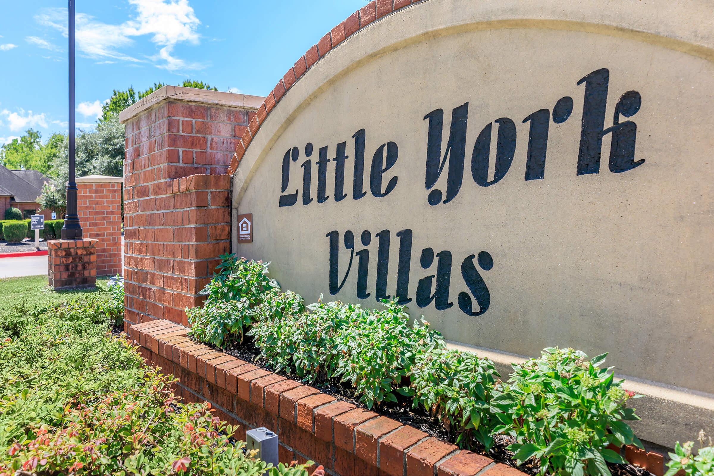 Sign for "Little York Villas" made of stone and brick, surrounded by greenery and colorful plants. The background features a clear blue sky with a few clouds, indicating a sunny day.