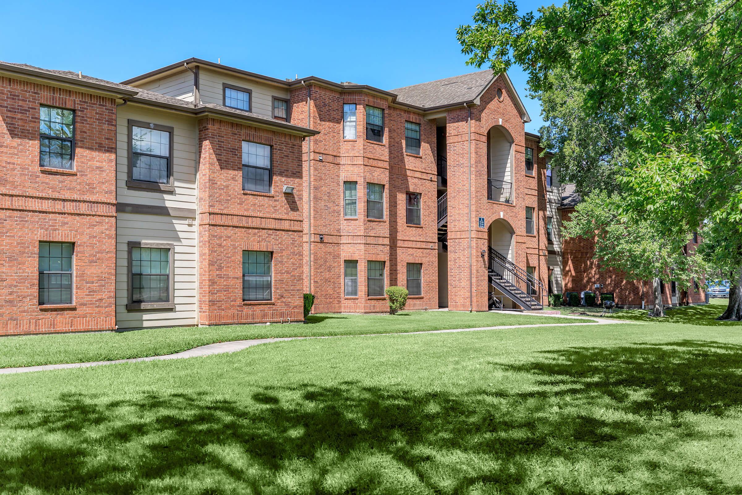 Brick apartment buildings surrounded by lush green grass and trees under a clear blue sky. The buildings feature multiple windows and a staircase leading to the entrance.