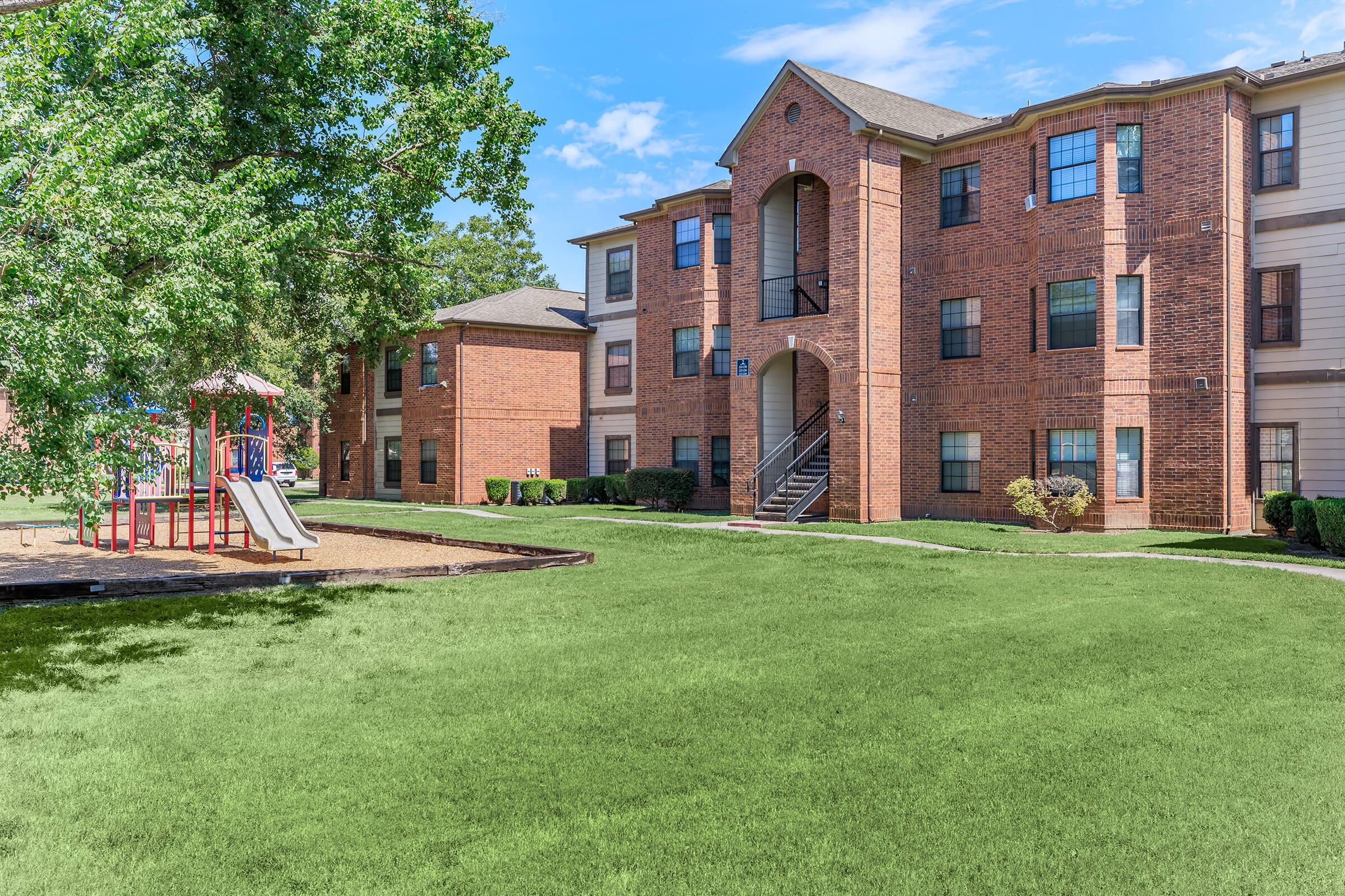 A well-maintained apartment complex with brick buildings, featuring a playground with a slide and swings in the foreground. Lush green grass surrounds the area, and trees provide shade. The setting is bright and inviting, suggesting a family-friendly environment.