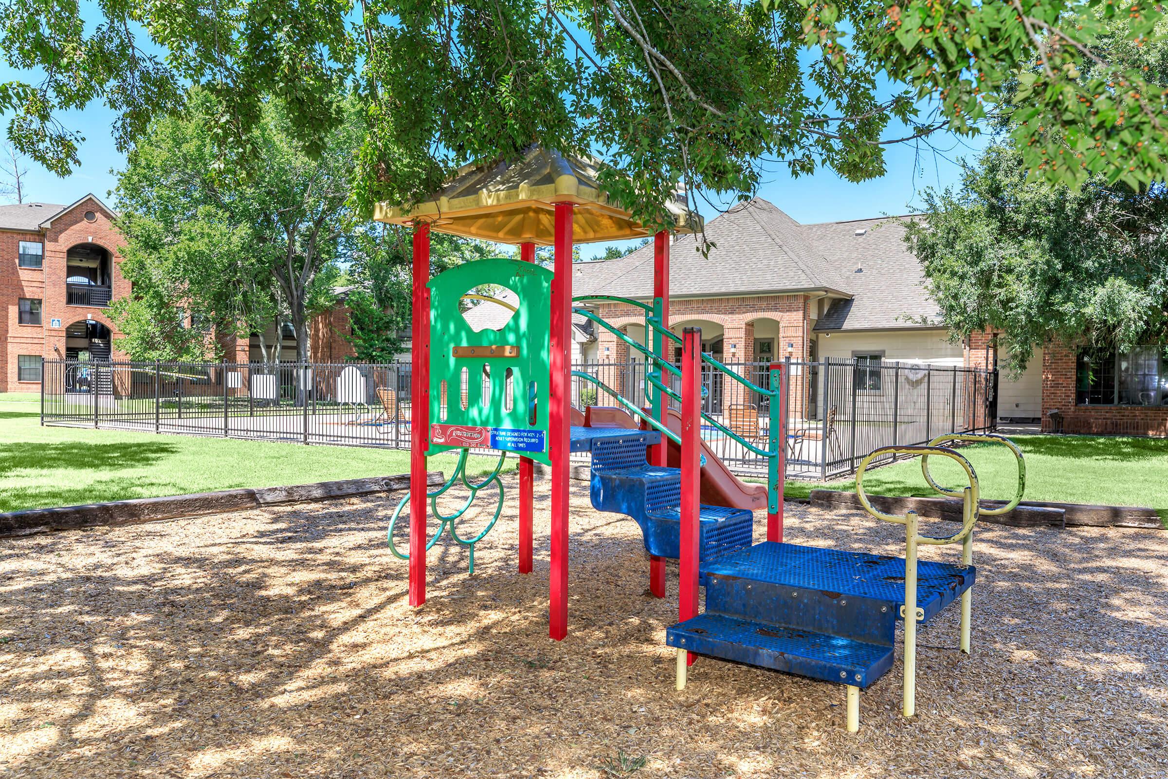 Colorful playground structure with a slide, climbing equipment, and swings, surrounded by a grassy area and shaded by trees. In the background, there are residential buildings and a fence enclosing the playground. The ground is covered with wood chips.
