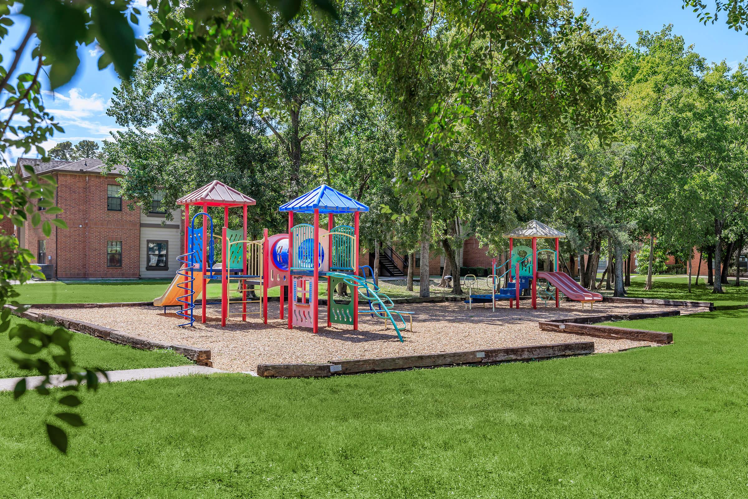 A colorful playground featuring multiple play structures, slides, and climbing equipment, surrounded by green grass and trees on a sunny day.