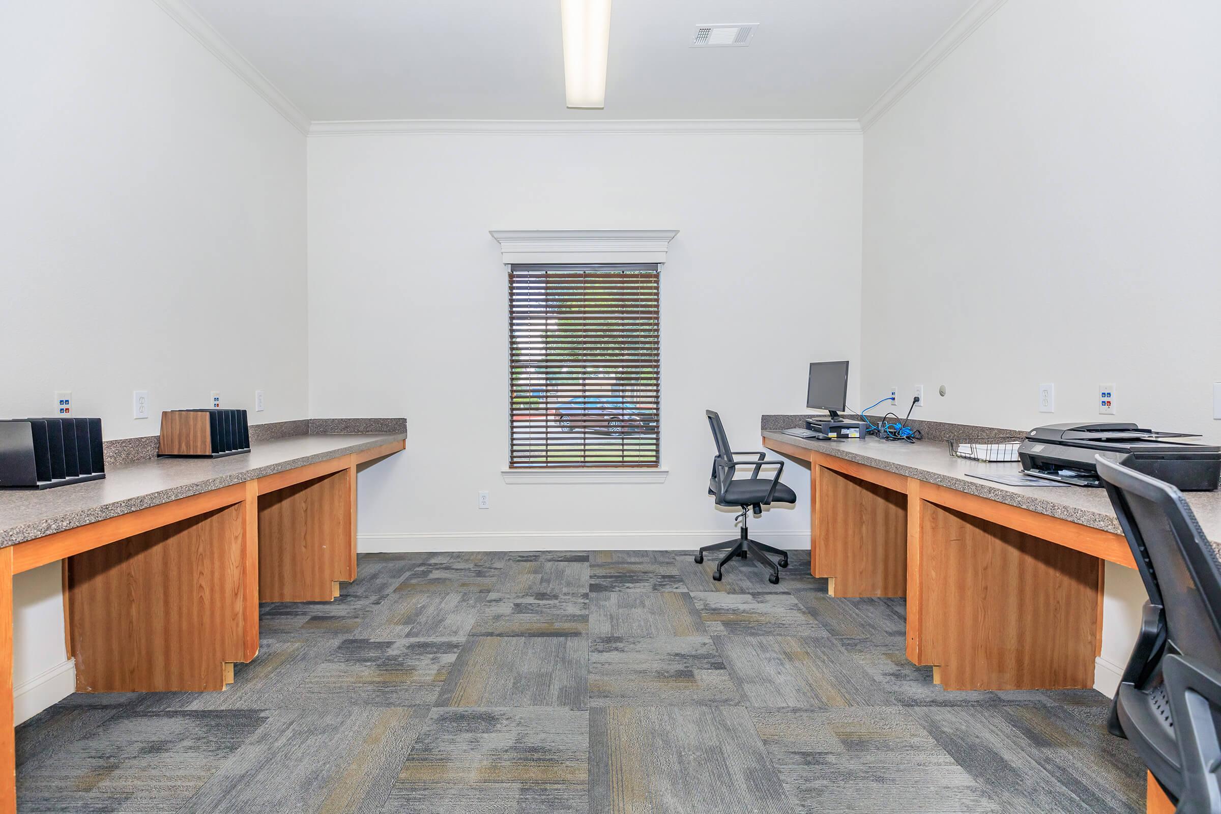 A spacious, modern office interior featuring two separate workstations with wooden desks and black chairs. The walls are painted white, and there are large windows with blinds letting in natural light. The floor is covered with grey and blue carpeting, creating a professional and organized atmosphere.