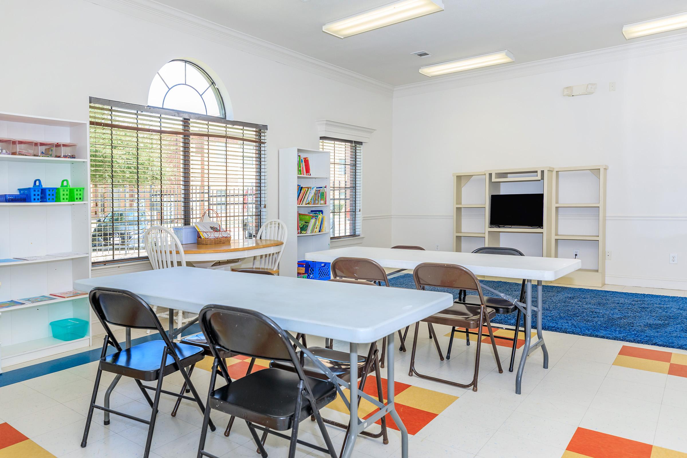 A bright and spacious classroom featuring several tables and chairs arranged for group activities. There are bookshelves with colorful blocks and books, a TV on a shelf, and large windows allowing natural light to fill the room. A blue rug is placed on the floor, creating a cozy atmosphere.