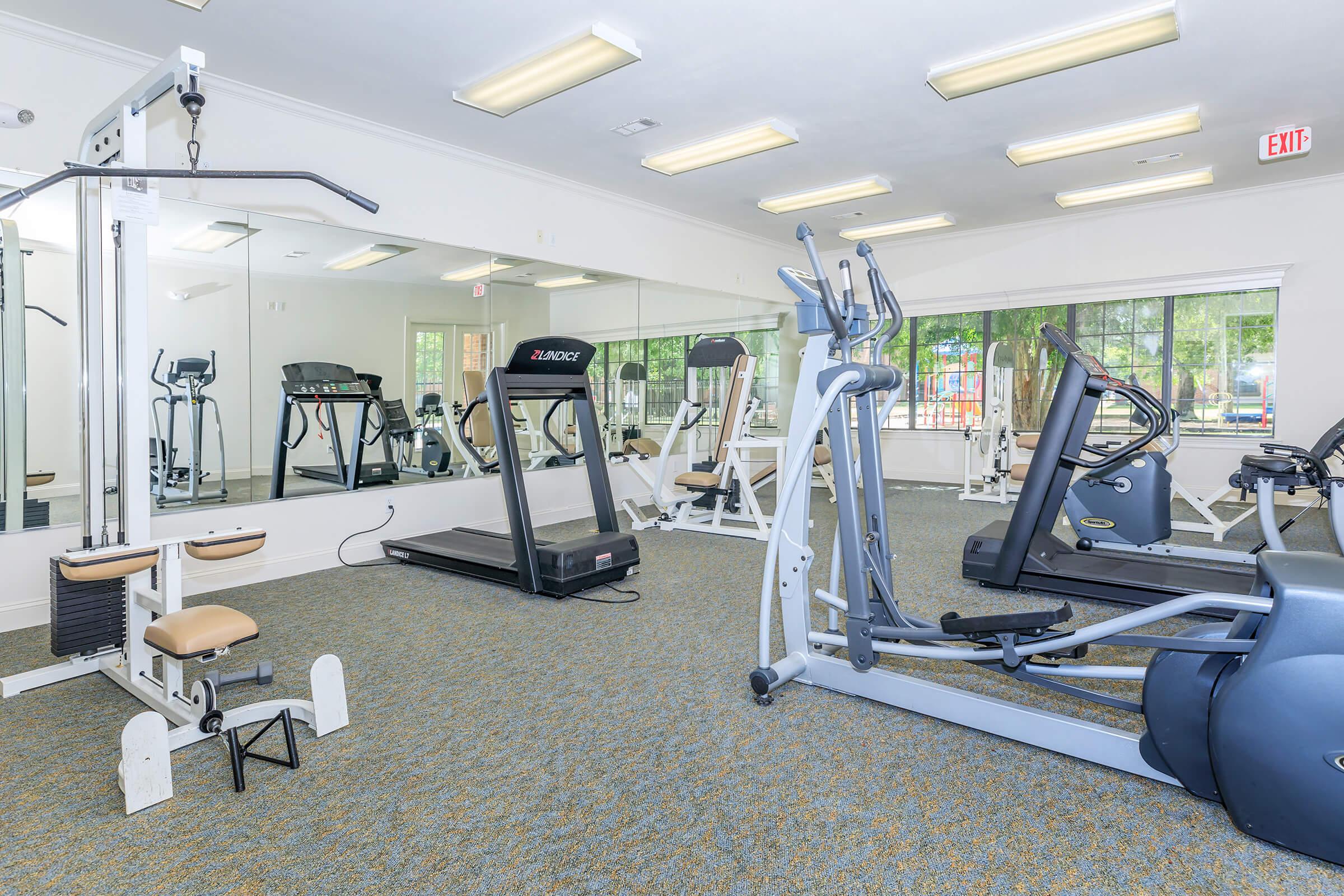 A well-equipped gym featuring various exercise machines, including a treadmill, elliptical, and strength training equipment. The space is bright and spacious, with large windows allowing natural light, and a clean carpeted floor.