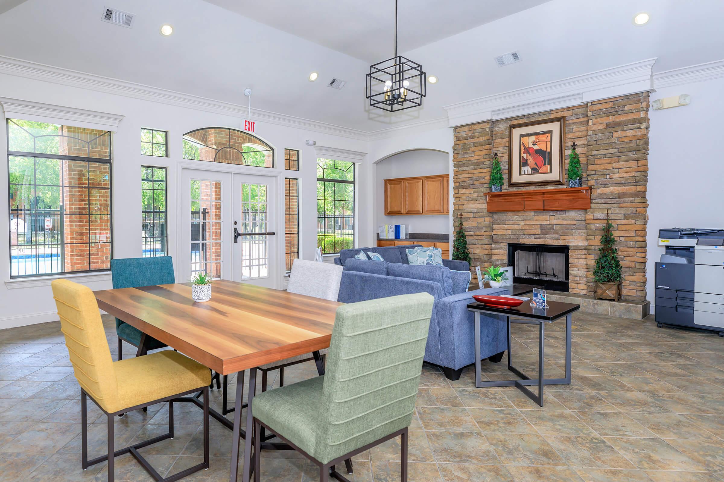 A modern communal lounge area featuring a wooden dining table surrounded by colorful chairs, a cozy blue sofa, and a stone fireplace. Large windows allow natural light in, and there are plants and a printer in the space, creating an inviting atmosphere for relaxation or meetings.