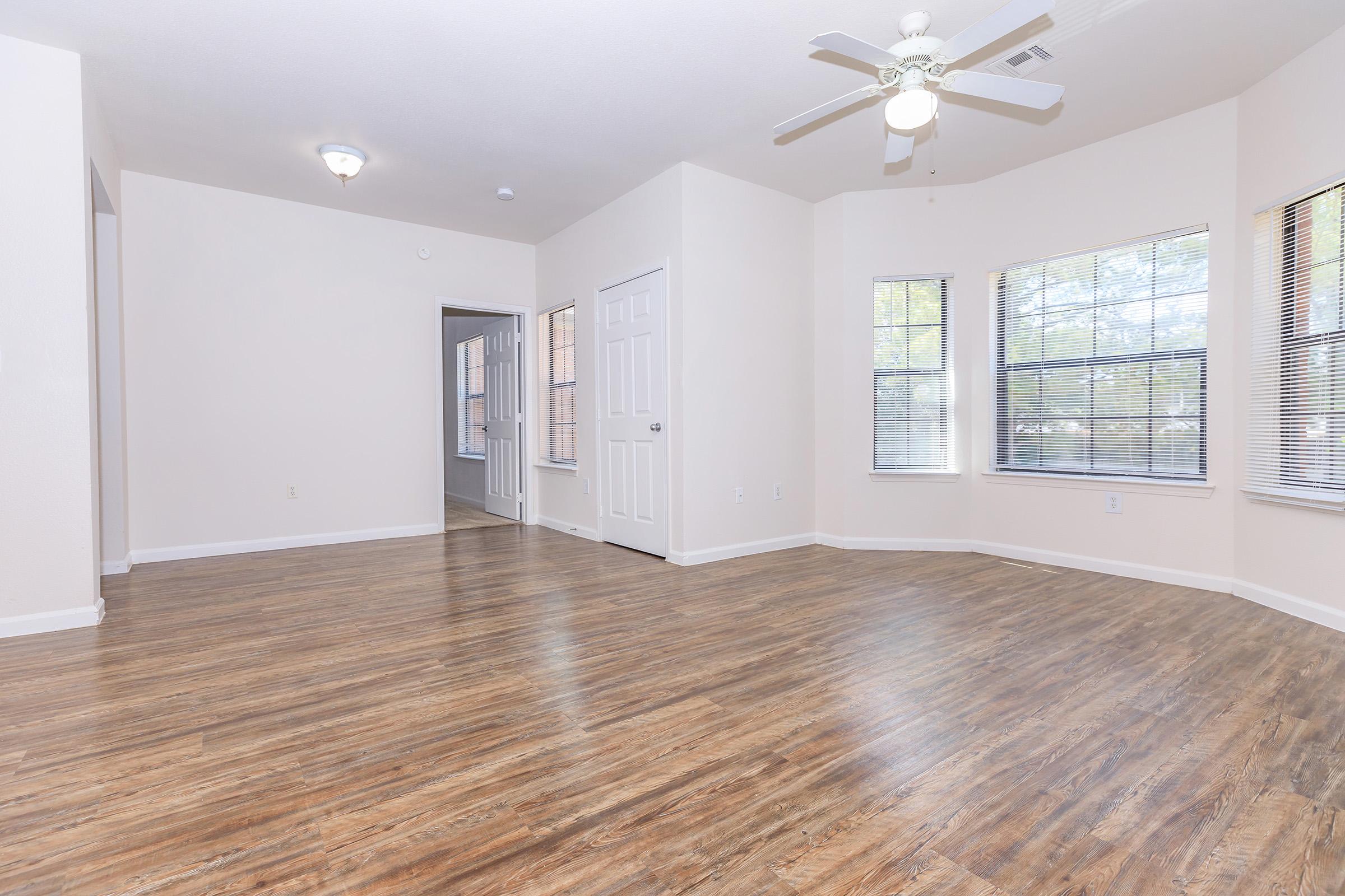 Spacious, well-lit living area featuring light wood flooring, a ceiling fan, and large windows. Doors lead to adjacent rooms, creating an open layout. The walls are painted in a neutral tone, enhancing the bright and airy atmosphere of the space.