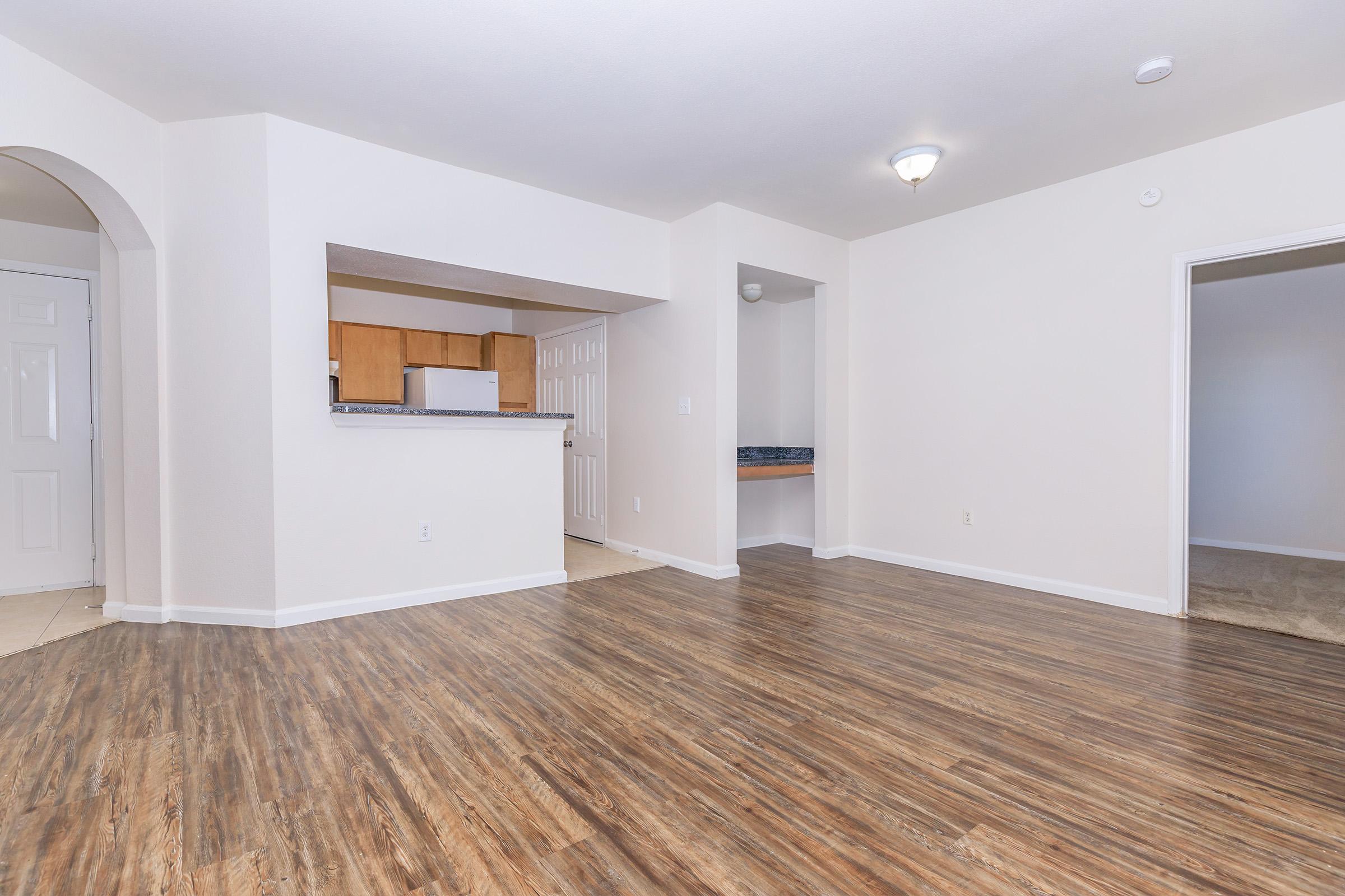 Spacious empty living area with wood-style flooring, arched entrance leading to a kitchen area with wooden cabinets, and an opening to a hallway. The walls are painted in a light color, providing a bright and airy atmosphere. The room is uncluttered and ready for furnishing.