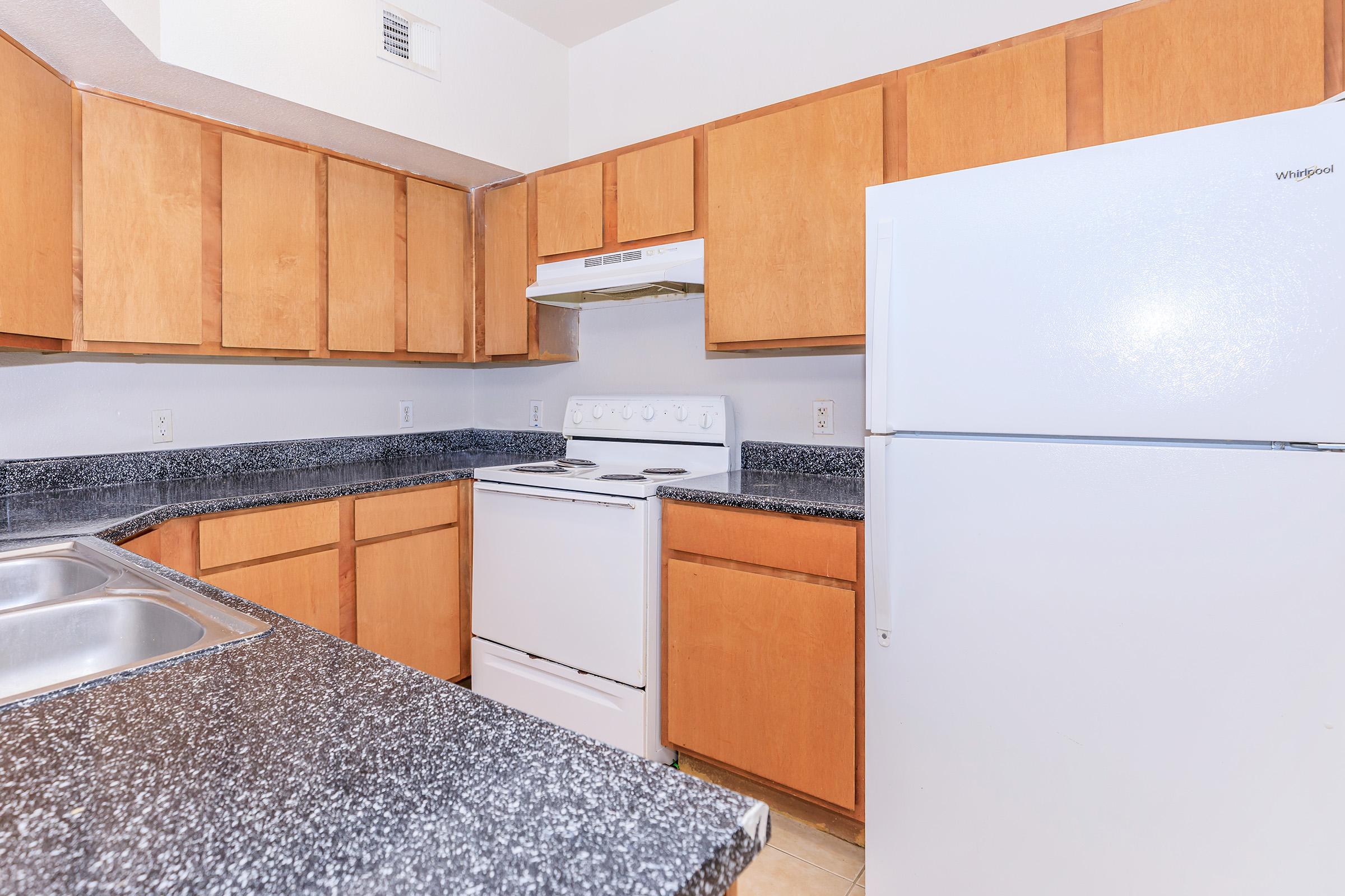 A modern kitchen featuring wooden cabinets, a white refrigerator, an electric stove with an oven, a silver kitchen sink, and dark countertop. The room is well-lit with a neutral color scheme, giving it an inviting and spacious look.
