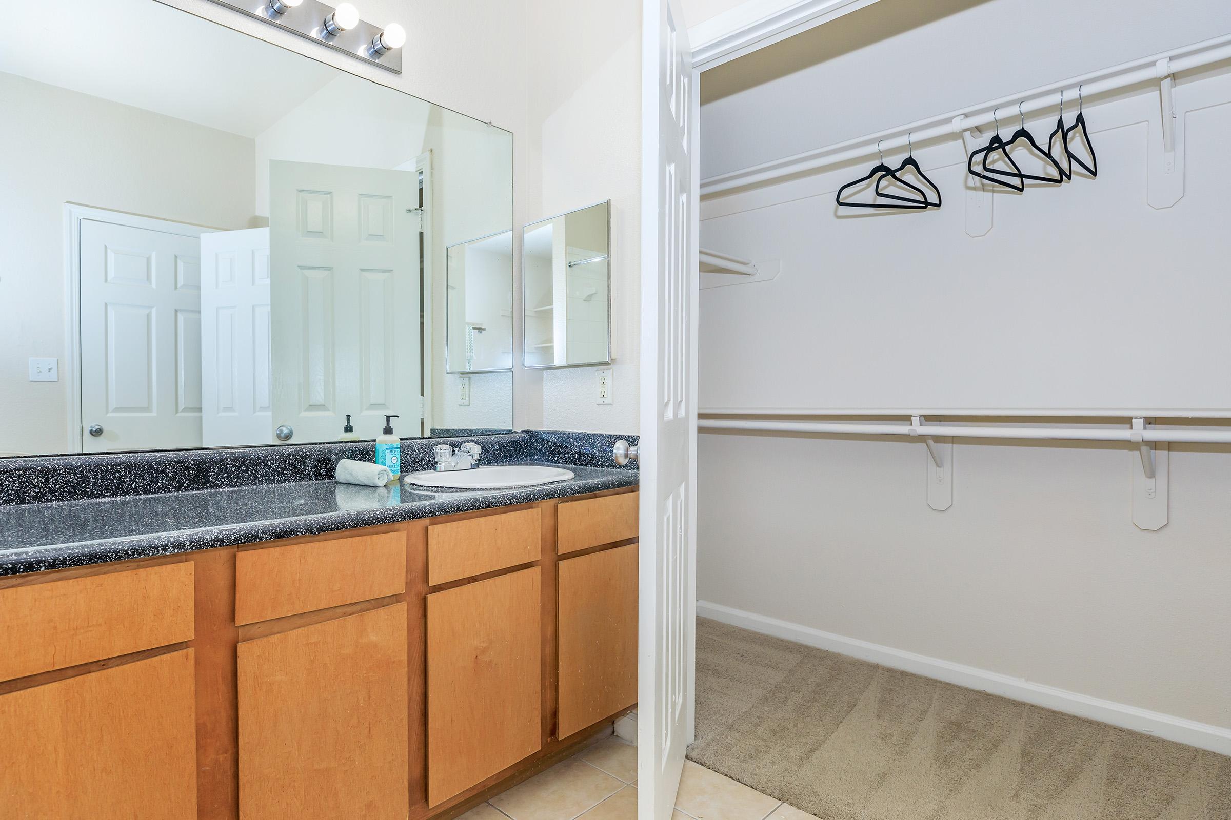 A well-lit bathroom featuring a double vanity with a black granite countertop, wooden cabinets, a large mirror, and a sink. To the right, an open closet with several hangers and a clean, neutral-colored carpet. The walls are light-colored, creating a spacious feel.