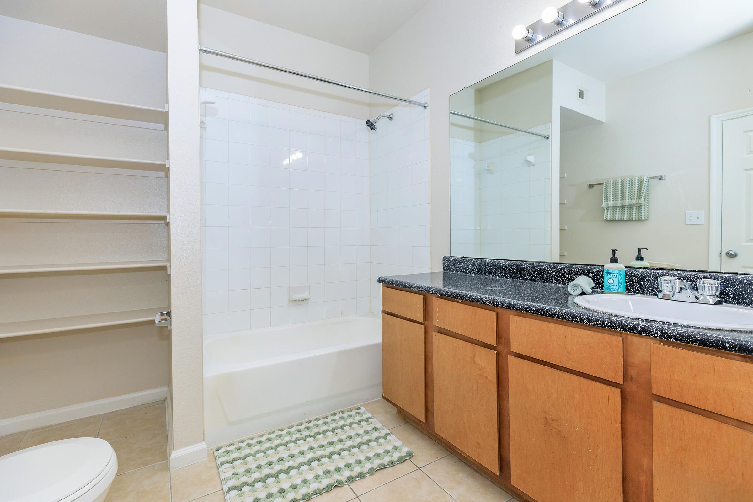 A clean, modern bathroom featuring a bathtub with a glass sliding door, a countertop with a sink and mirror, and light-colored walls. There are open shelves for storage and a patterned rug in front of the bathtub. Bright lighting above the mirror adds to the spacious feel.