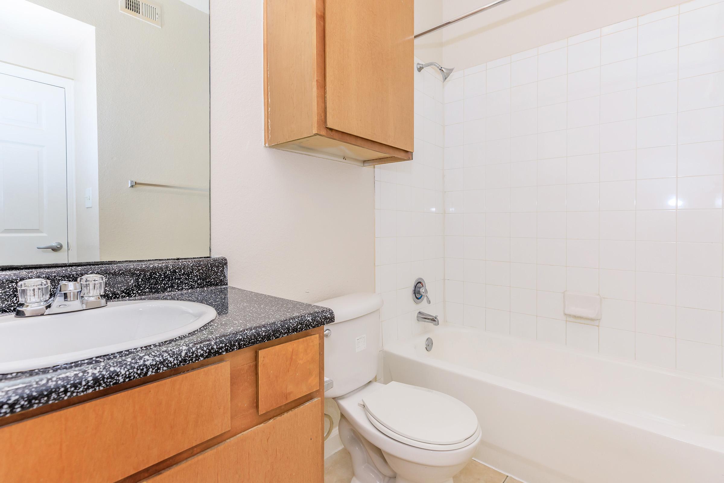 A clean and simple bathroom featuring a white bathtub with a shower, a toilet, and a sink with a dark countertop. Light-colored walls and a large mirror enhance the space. The bathroom is well-lit, with neutral tiles on the floor and cabinetry providing storage.