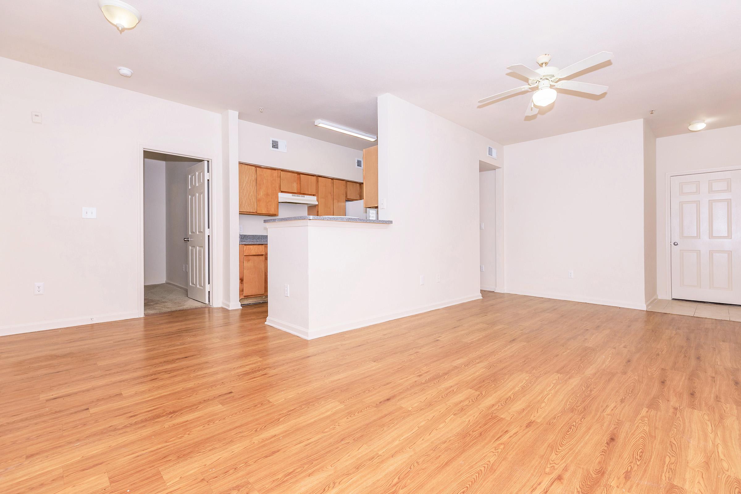 Spacious, unfurnished living area with light hardwood flooring, a ceiling fan, and an open layout. A doorway leads to a kitchen with wooden cabinets, and there's a door nearby opening to a hallway. The walls are painted in light colors, enhancing the bright and airy feel of the space.