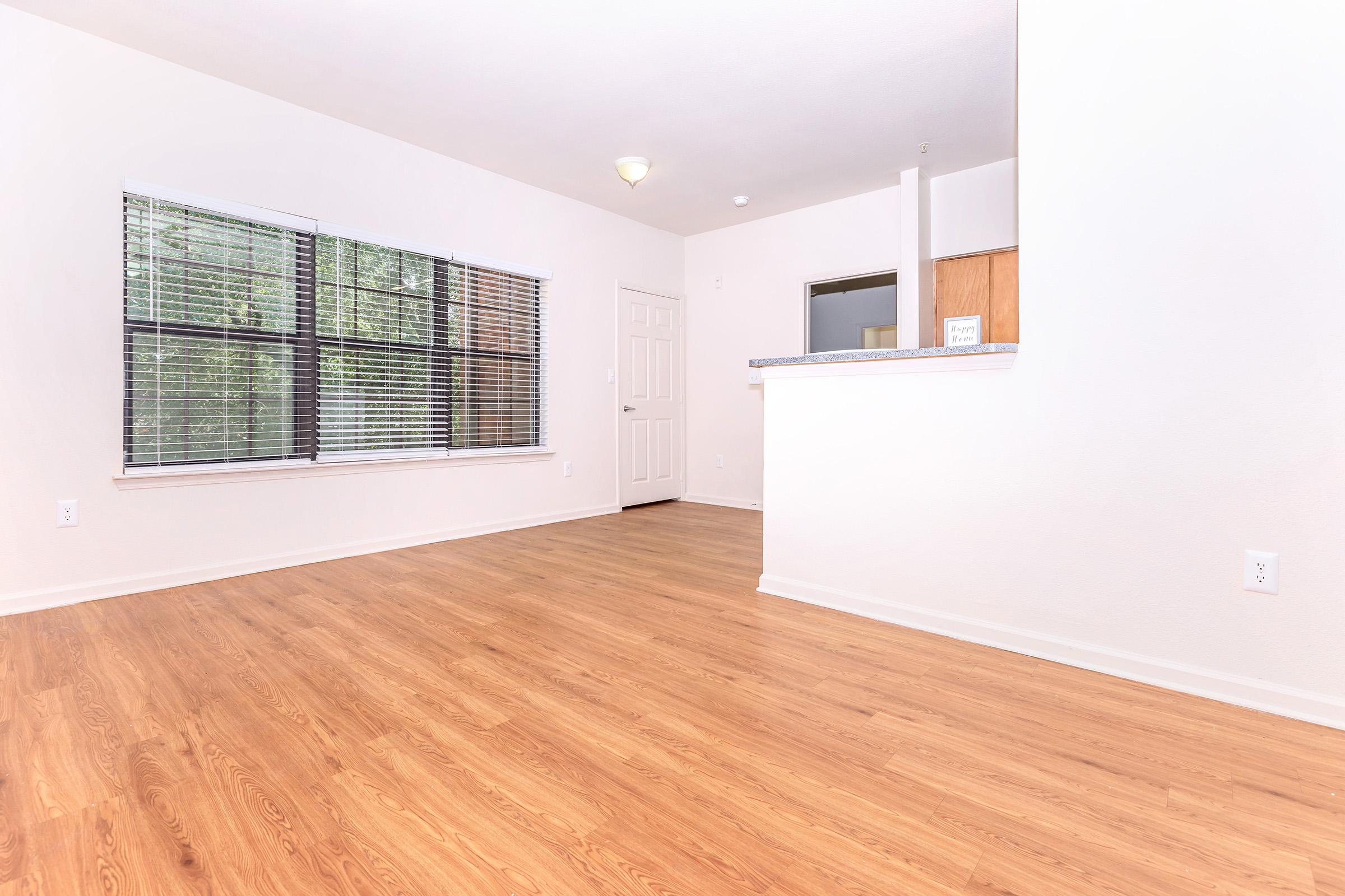 Spacious living area with hardwood floors, large window with blinds, and a doorway leading to a kitchen bar area. The walls are painted in neutral tones, creating a bright and inviting atmosphere.