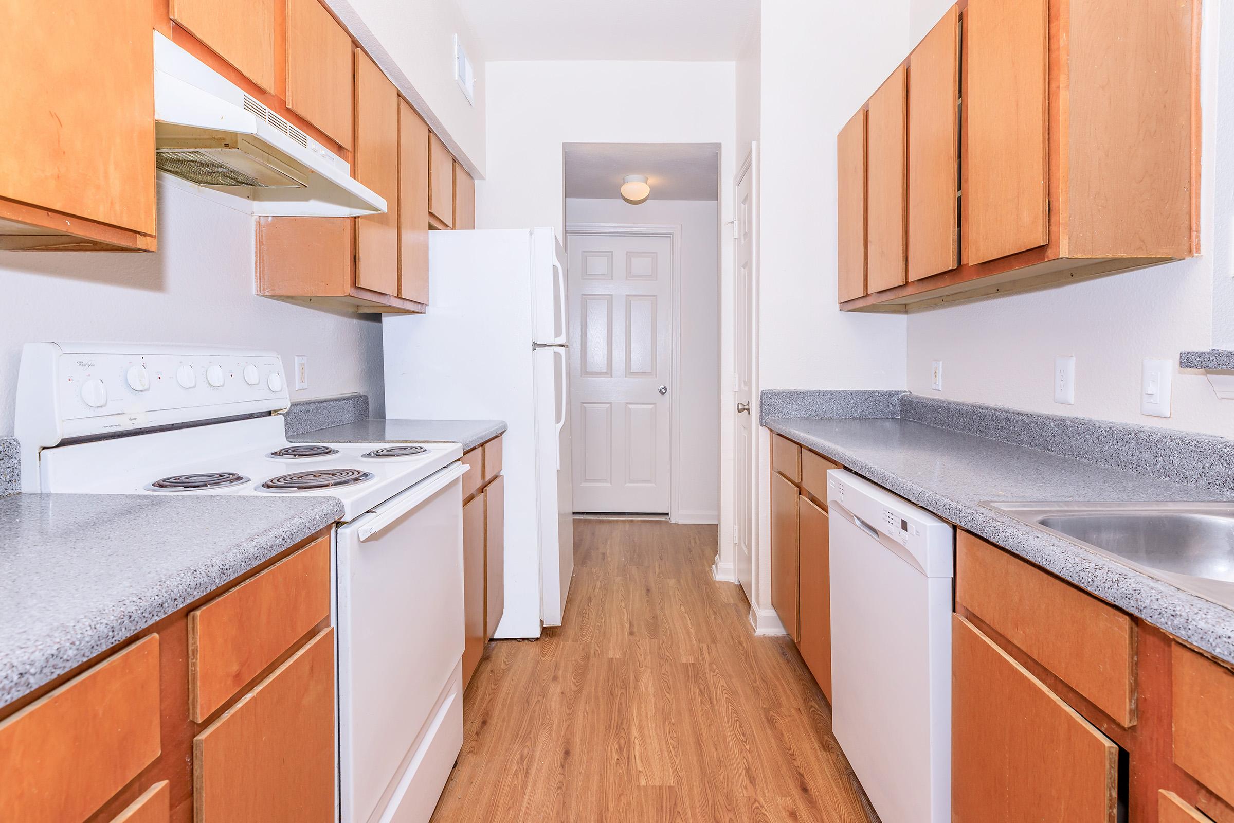 A modern kitchen featuring wooden cabinets, a white stove, dishwasher, and refrigerator. The countertops are gray, and the flooring is light wood. The kitchen is well-lit with a doorway leading to another room at the end.