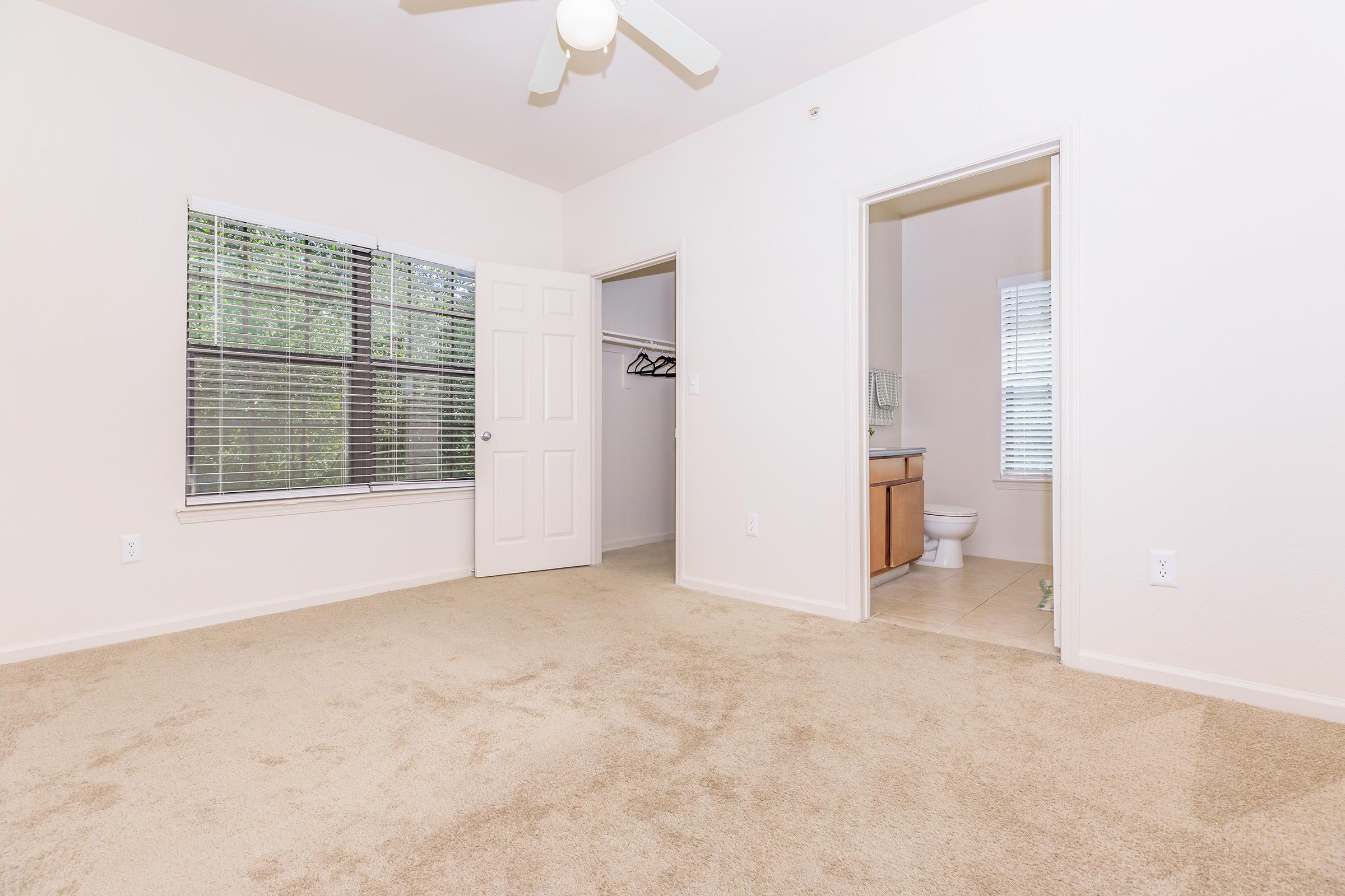 A spacious, neutral-colored empty bedroom featuring a ceiling fan, carpeted flooring, and two doors leading to adjacent areas. One door opens to a closet with hangers, while the other leads to a bathroom with natural light from a nearby window.