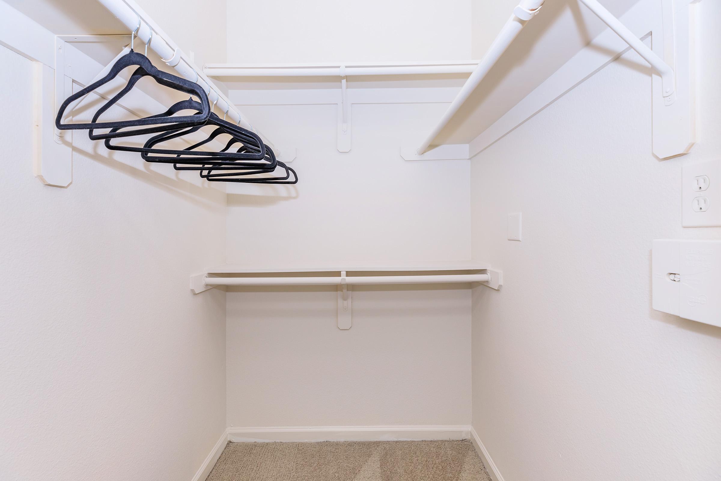 A small, empty closet featuring a light-colored wall, a carpeted floor, and several white hanging rods. Black plastic hangers are neatly hung on one rod, while an empty shelf is visible above. The space appears organized and uncluttered, ideal for storing clothes and accessories.