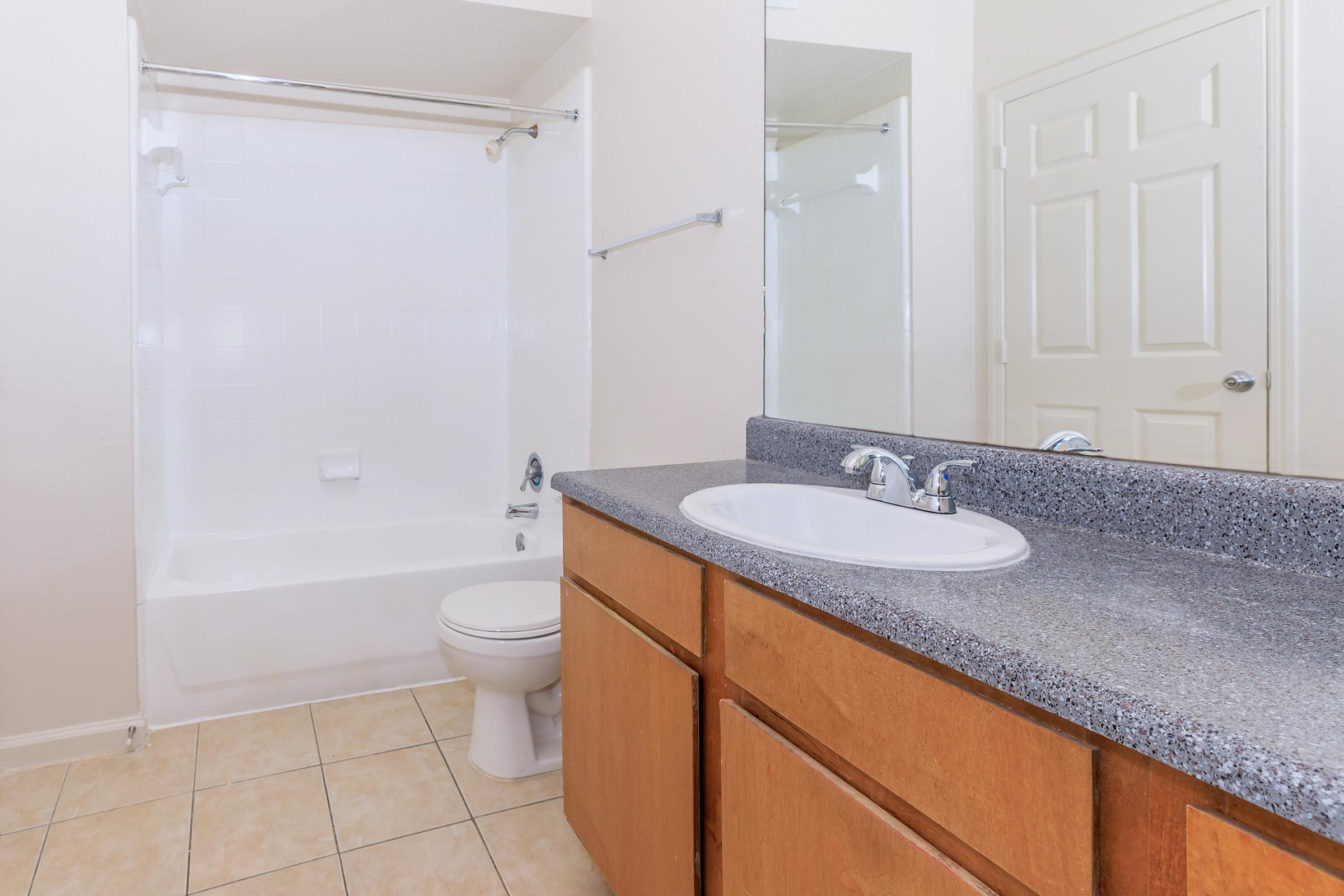 A clean bathroom featuring a white bathtub with a shower, a single sink vanity with granite countertops, wooden cabinets, and tiled flooring. A mirror above the sink and a closed toilet are also visible, along with beige walls and a door in the background.