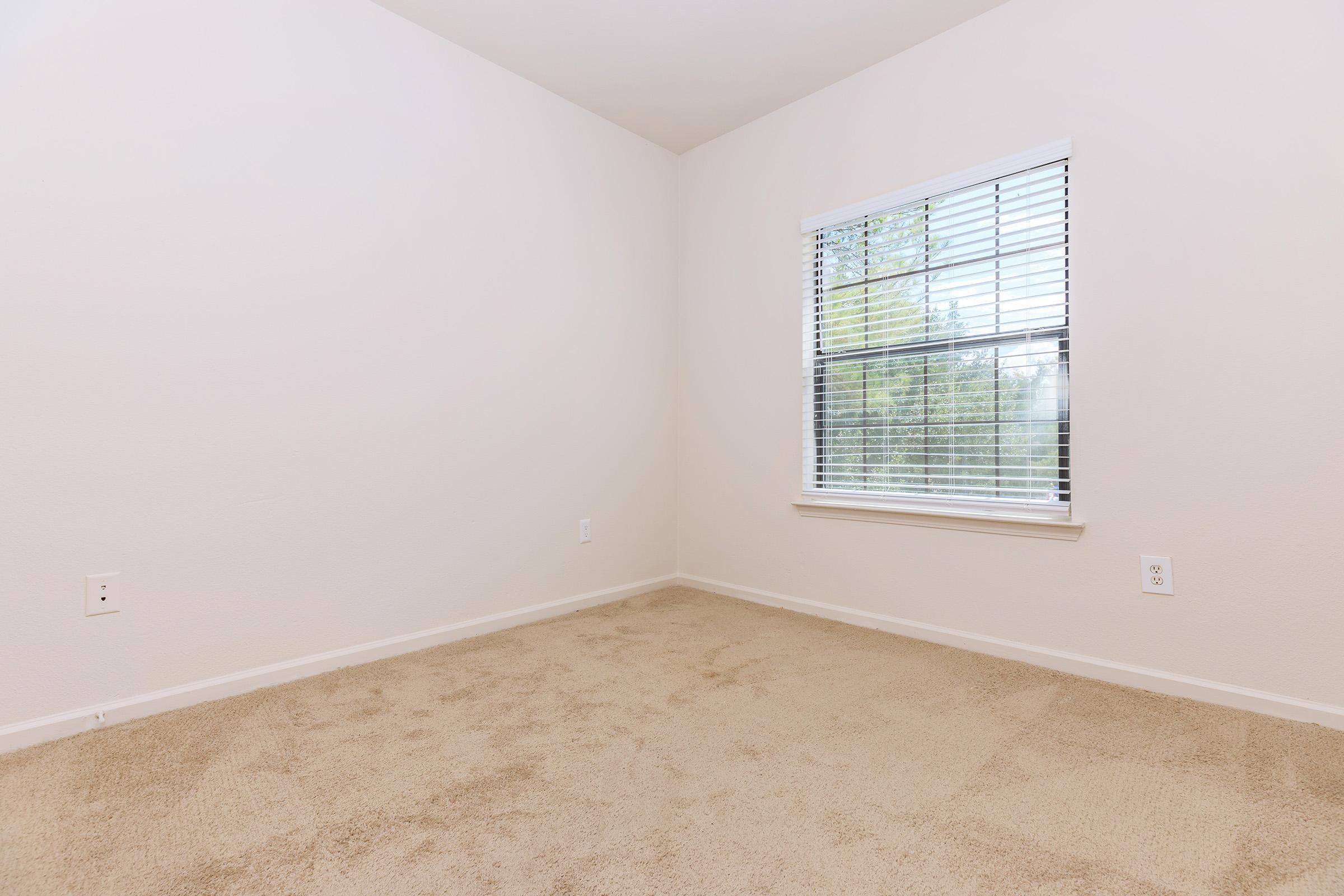 A clean, empty room with light beige walls and carpet. There is a window with white blinds allowing natural light, and no furniture or decorations are present. The floor is carpeted, creating a neutral and spacious atmosphere.