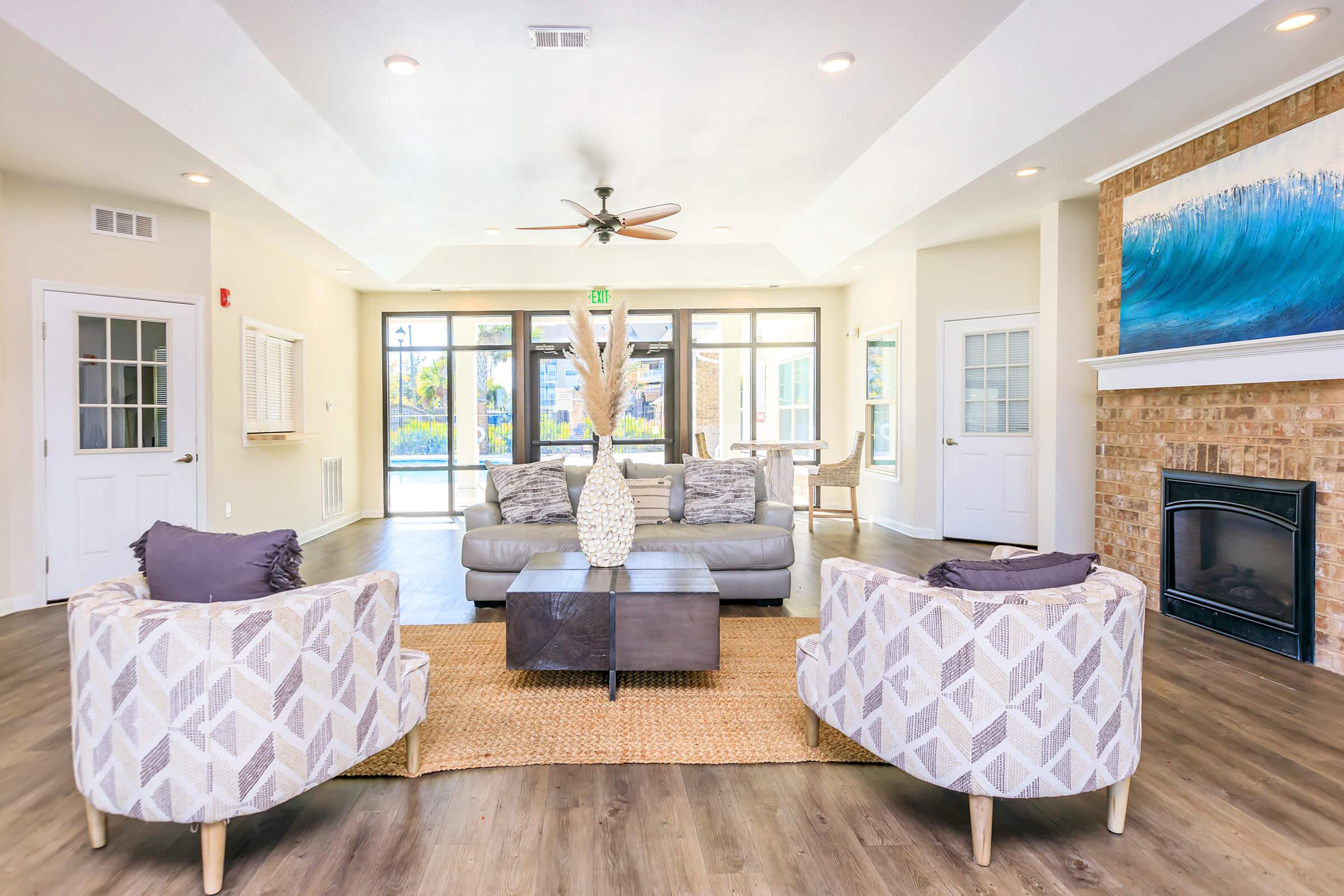 A bright and modern living room featuring a comfortable gray sofa and two patterned armchairs. A coffee table sits in the center, and a large piece of artwork with blue waves is displayed on the fireplace wall. Natural light fills the space through large windows, with a cozy atmosphere created by wooden flooring.