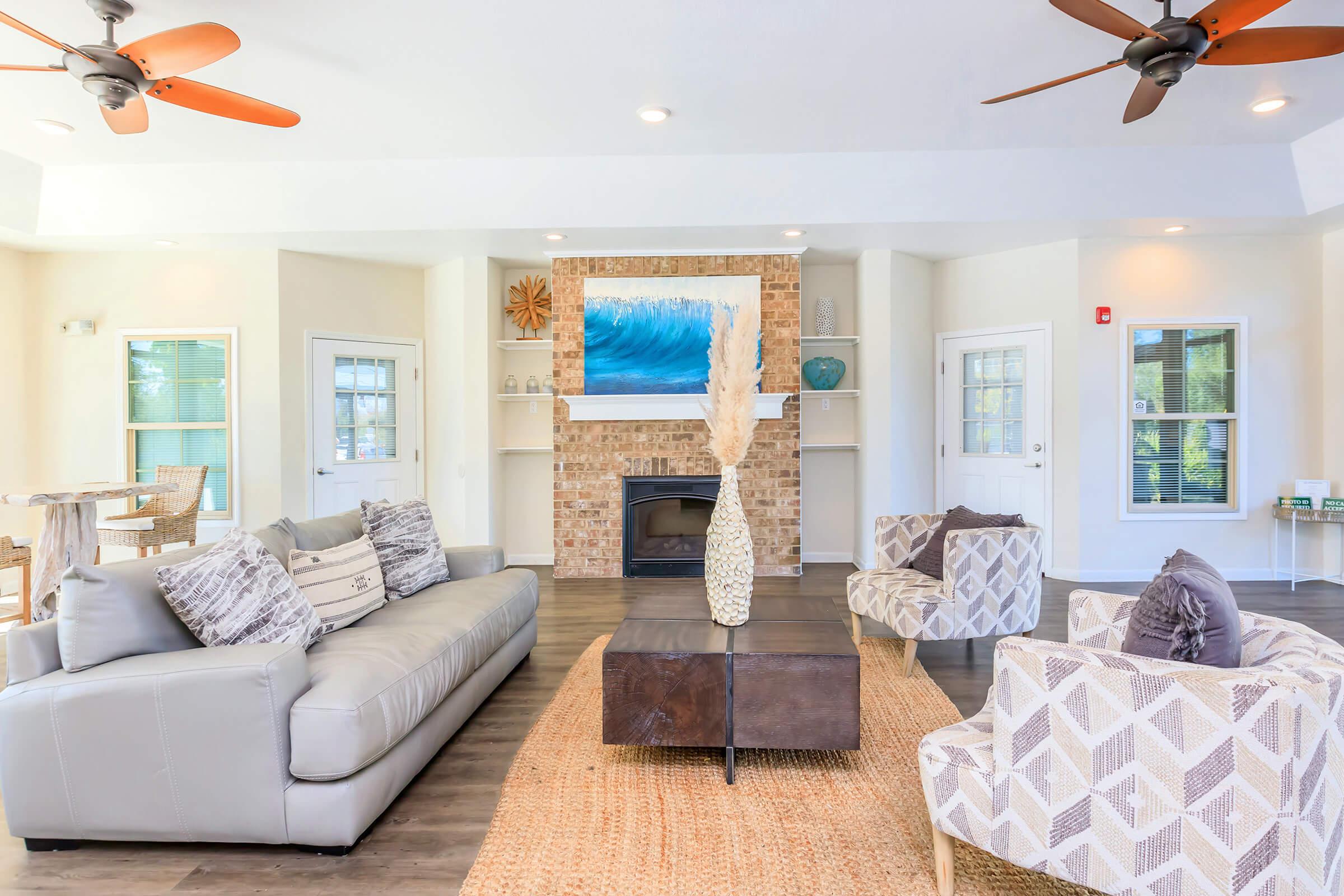 A bright and modern living room featuring a light gray sofa and patterned armchairs. A coffee table sits on a woven rug in the center. The walls are painted in soft tones, with a large piece of artwork depicting ocean waves above a brick fireplace. Two ceiling fans are visible, enhancing the airy feel of the space.