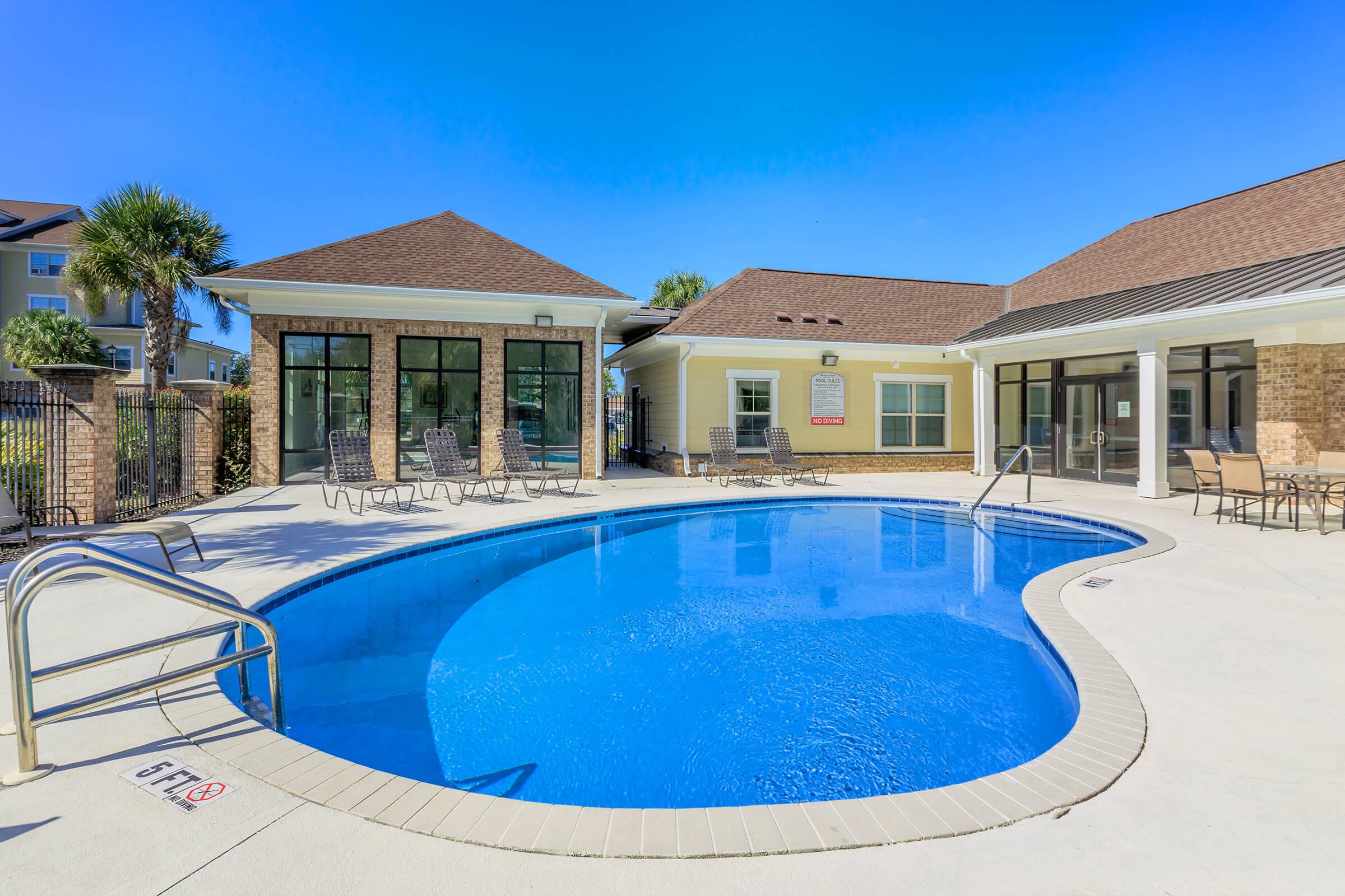 A sparkling blue swimming pool surrounded by a sunlit deck with lounge chairs. In the background, there are two buildings with a modern design, featuring large windows and a tropical landscaping. Clear skies enhance the inviting atmosphere of the pool area.