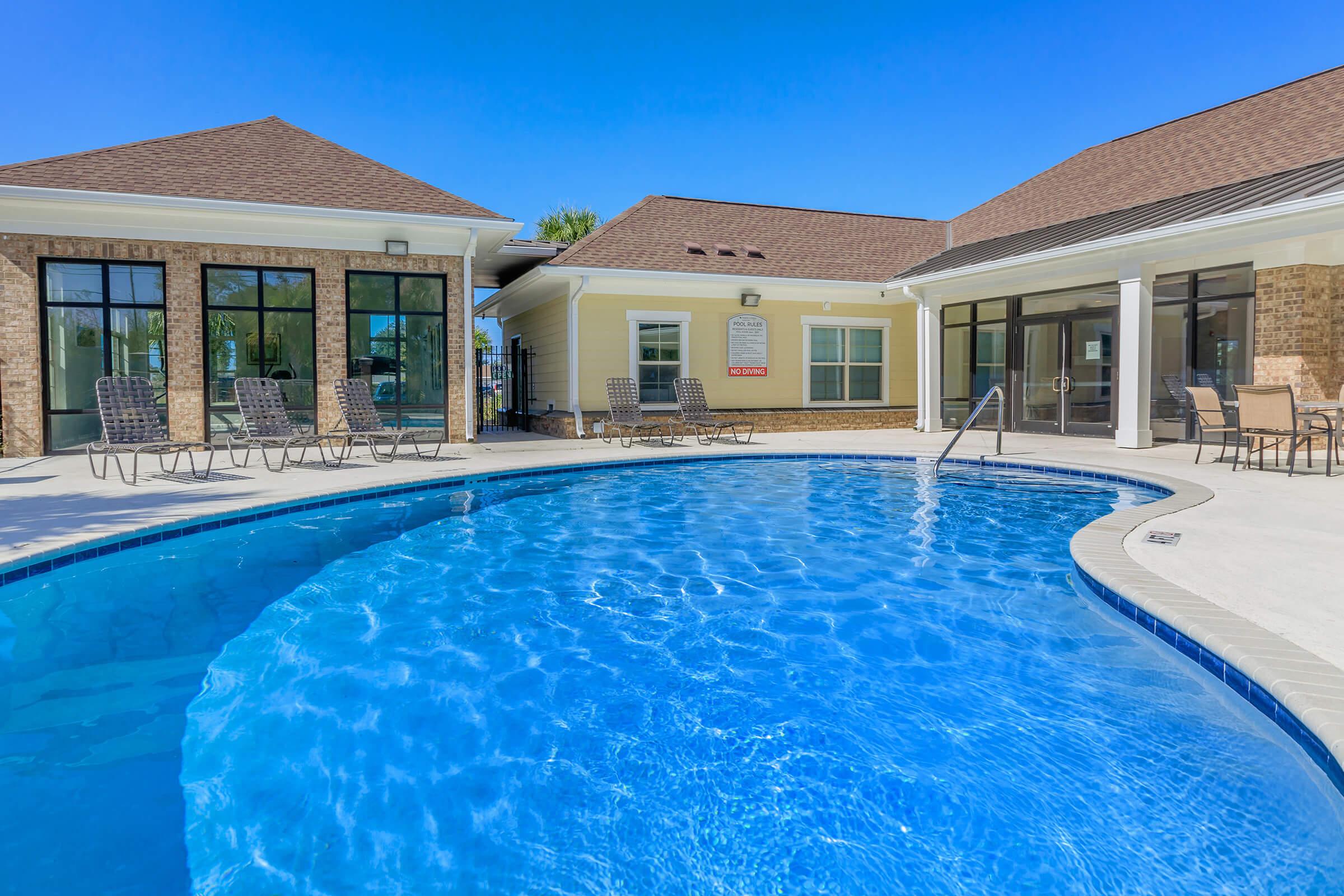 A clear blue swimming pool surrounded by a tan-colored patio. Comfortable lounge chairs are placed around the pool area. In the background, there are two buildings with large windows and a sign visible on one wall. The sky above is bright blue, indicating a sunny day.