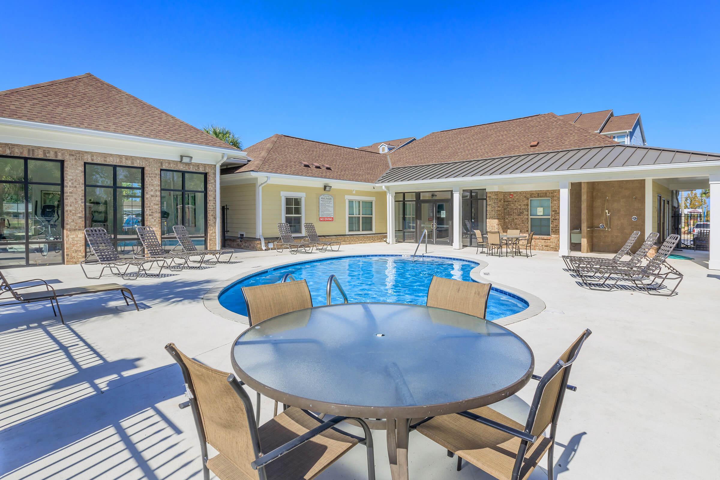 A clear blue sky overlooks a residential pool area featuring a circular pool surrounded by lounge chairs. A patio table with chairs sits in the foreground. The building has a mix of stone and light-colored siding, creating a welcoming outdoor space for relaxation and leisure activities.