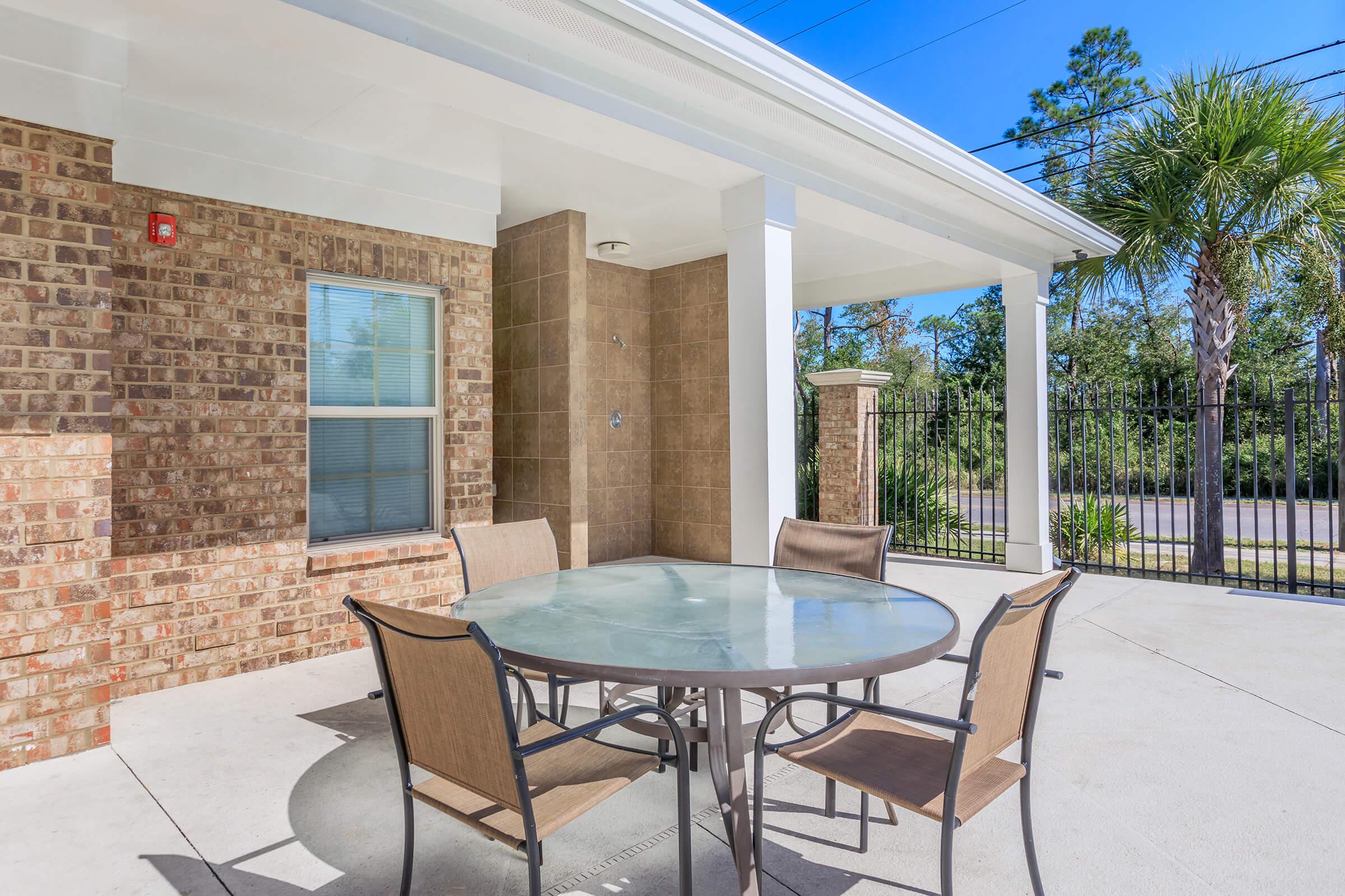 Outdoor patio area featuring a circular glass-top table surrounded by four beige chairs. The backdrop includes a brick wall and a tall wooden fence, with greenery visible in the distance. Bright, sunny weather is highlighted by a clear blue sky.