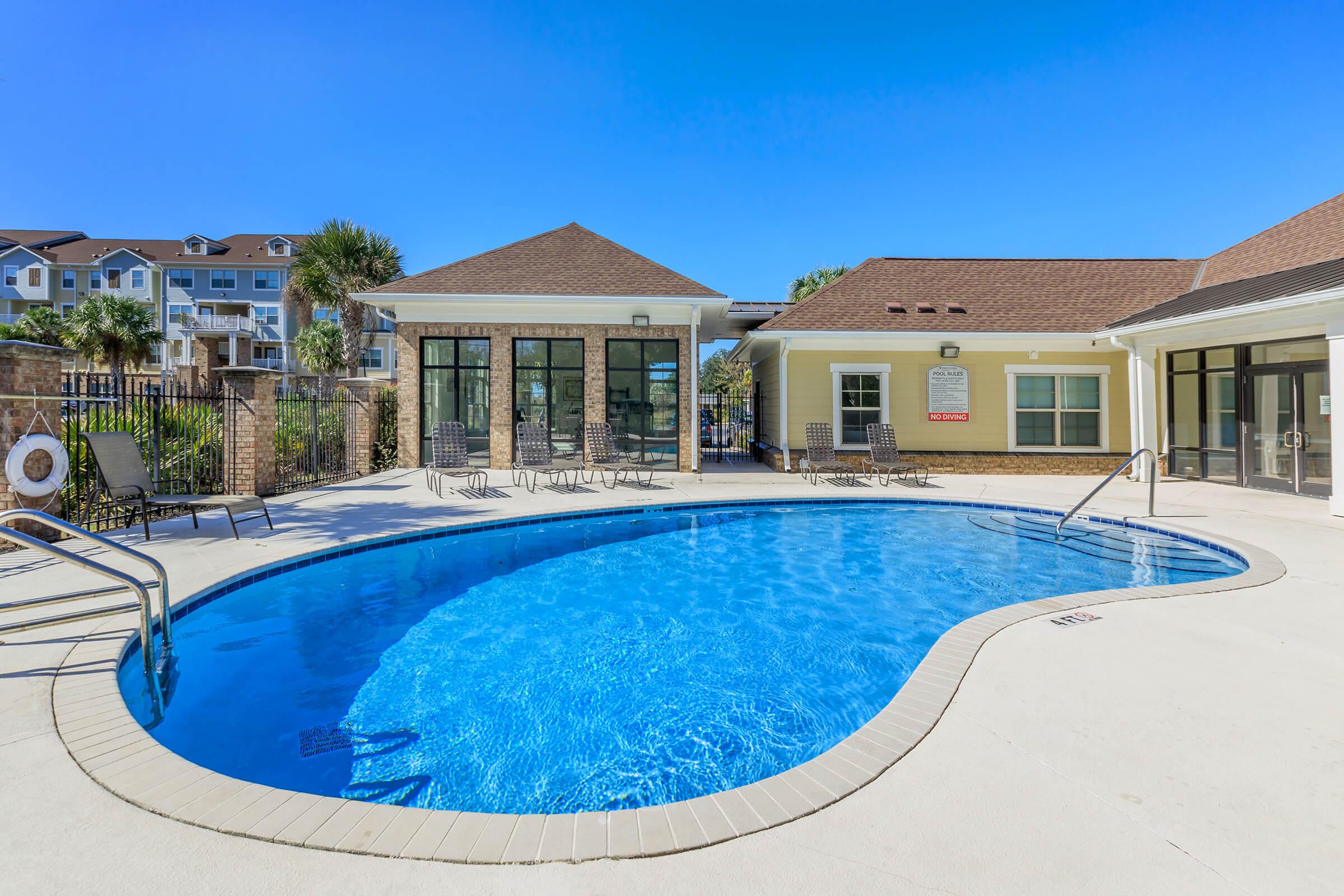 A sunny outdoor pool area featuring a curved blue pool with a ramp, surrounded by lounge chairs. In the background, there are two buildings with large windows and a clear blue sky overhead. The space is well-maintained and inviting for leisure activities.