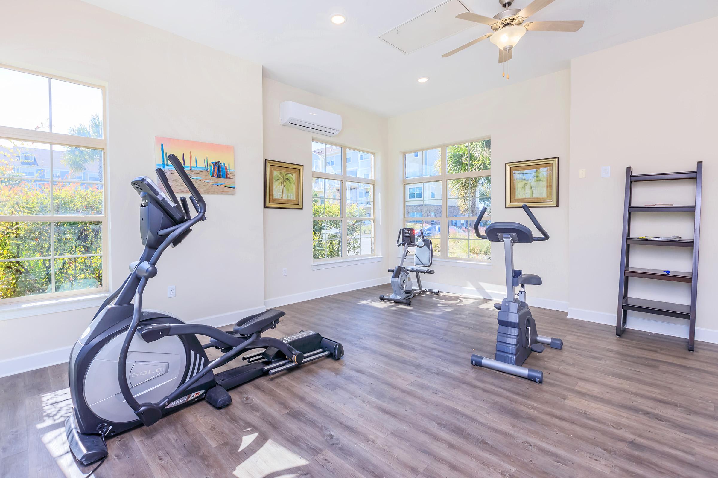 A bright and spacious fitness room featuring exercise equipment including an elliptical machine and a stationary bike, with large windows allowing natural light, wooden flooring, and wall art. A shelf is against one wall, enhancing the inviting atmosphere of the workout space.