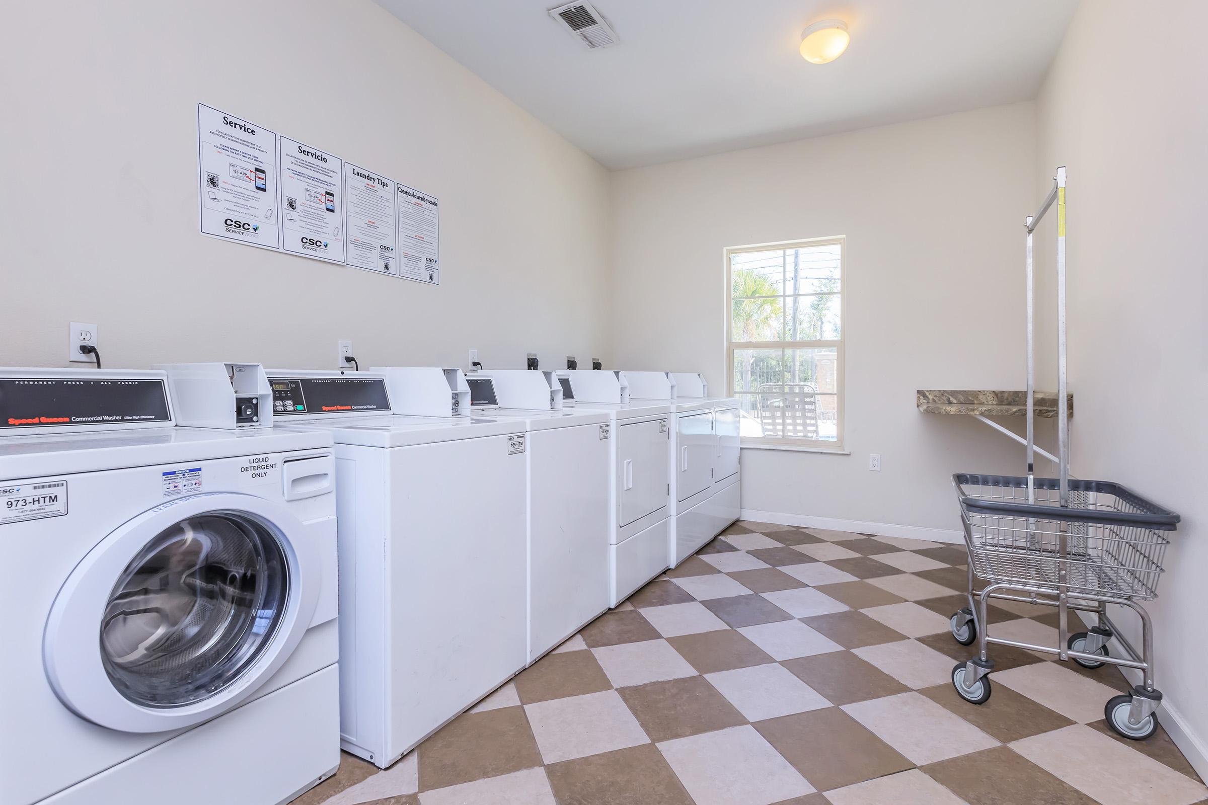 A clean laundry room featuring several white washing machines and dryers aligned against a wall. The floor has a checkered pattern of light and dark tiles. A window lets in natural light, and there is a folding table and a laundry cart nearby. Wall-mounted instructions for machine use are visible above the machines.