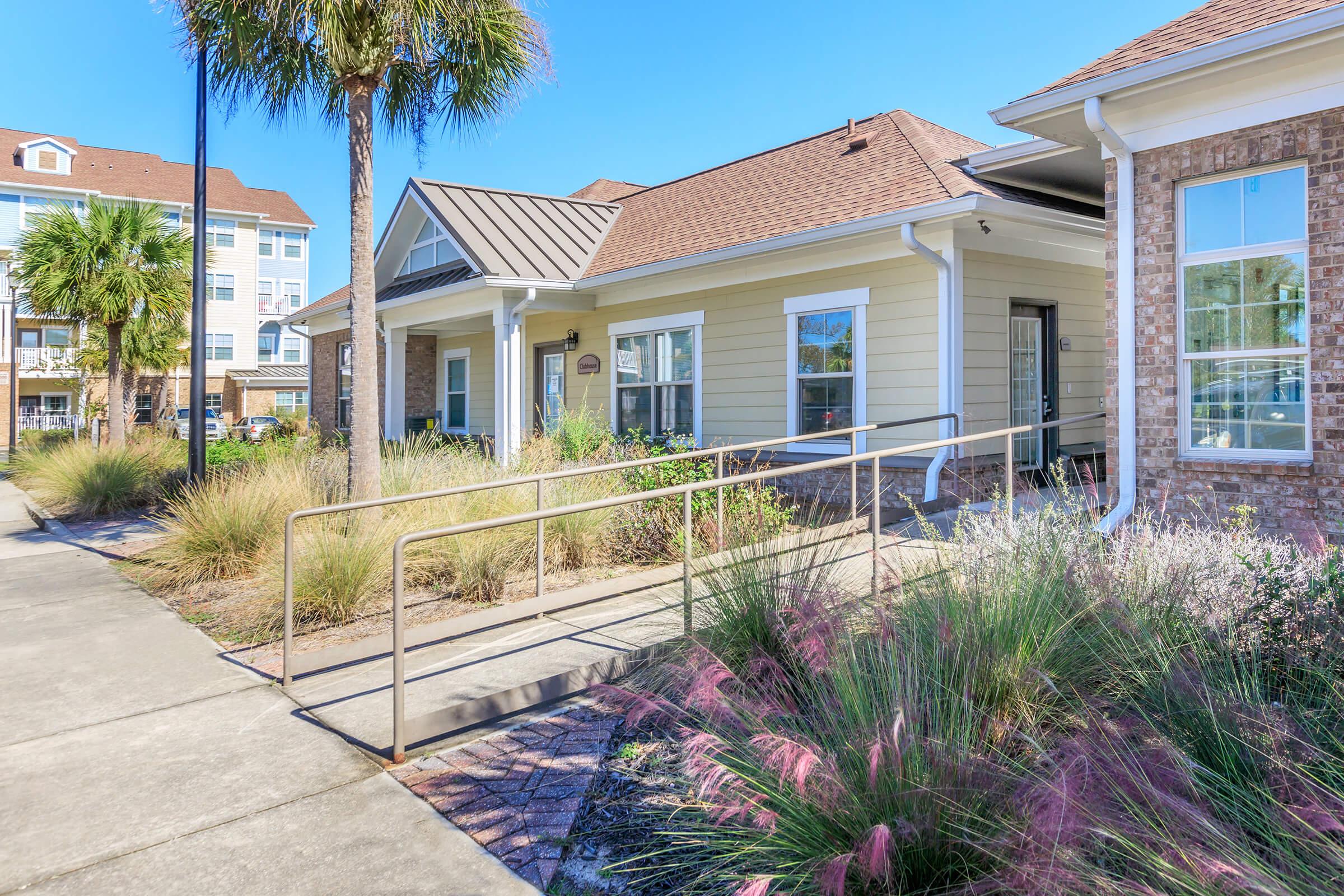 A modern building featuring a ramp for accessibility, surrounded by low landscaping with various plants. Palm trees are visible in the background, along with a multi-storied apartment community. The sky is clear and sunny, highlighting the welcoming design of the entrance.