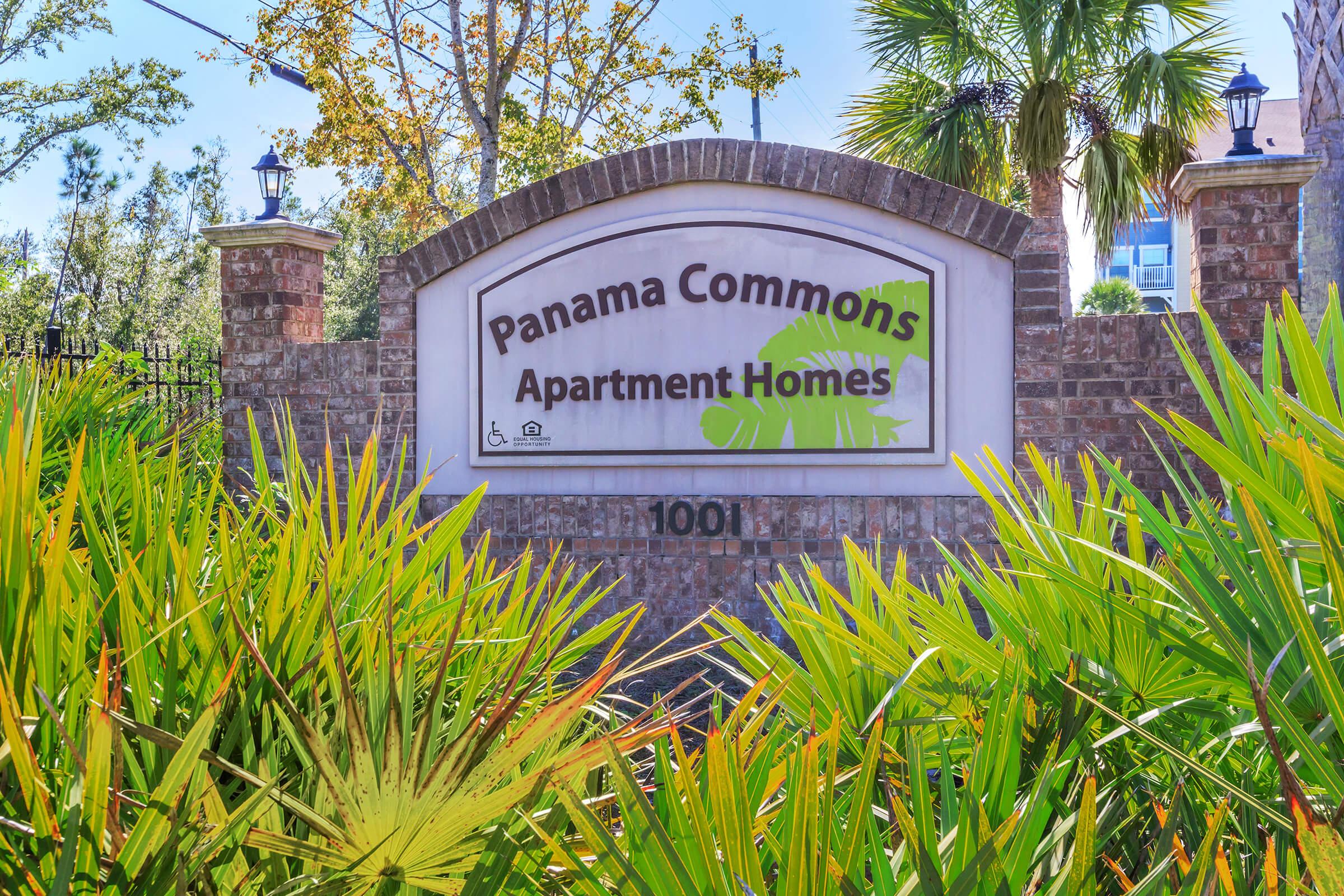 Sign for Panama Commons Apartment Homes, featuring the name prominently displayed. The sign is surrounded by vibrant green foliage and palm trees, with brick pillars on either side. The address "1001" is visible beneath the main title.