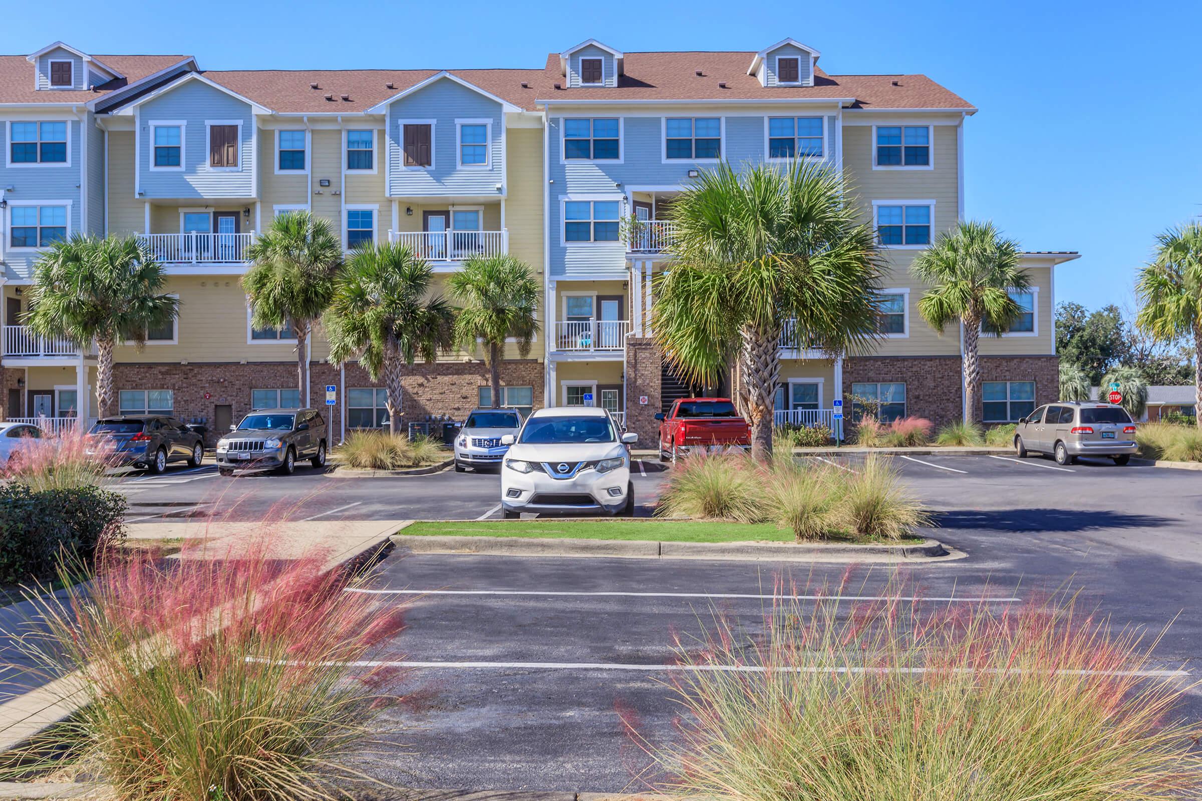 A sunny residential complex featuring multiple floors with balconies, surrounded by palm trees and landscaped grass. Several cars are parked in the foreground, and the building's exterior is a mix of light colors, showcasing a welcoming atmosphere.