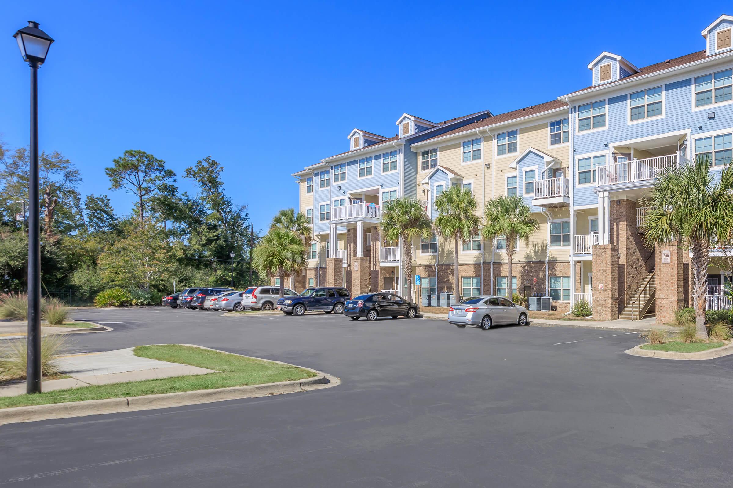 A multi-story apartment building with balconies and palm trees in front. The parking lot is partially filled with cars, and there are street lamps illuminating the area. The sky is clear and blue, suggesting a sunny day. Trees can be seen in the background.