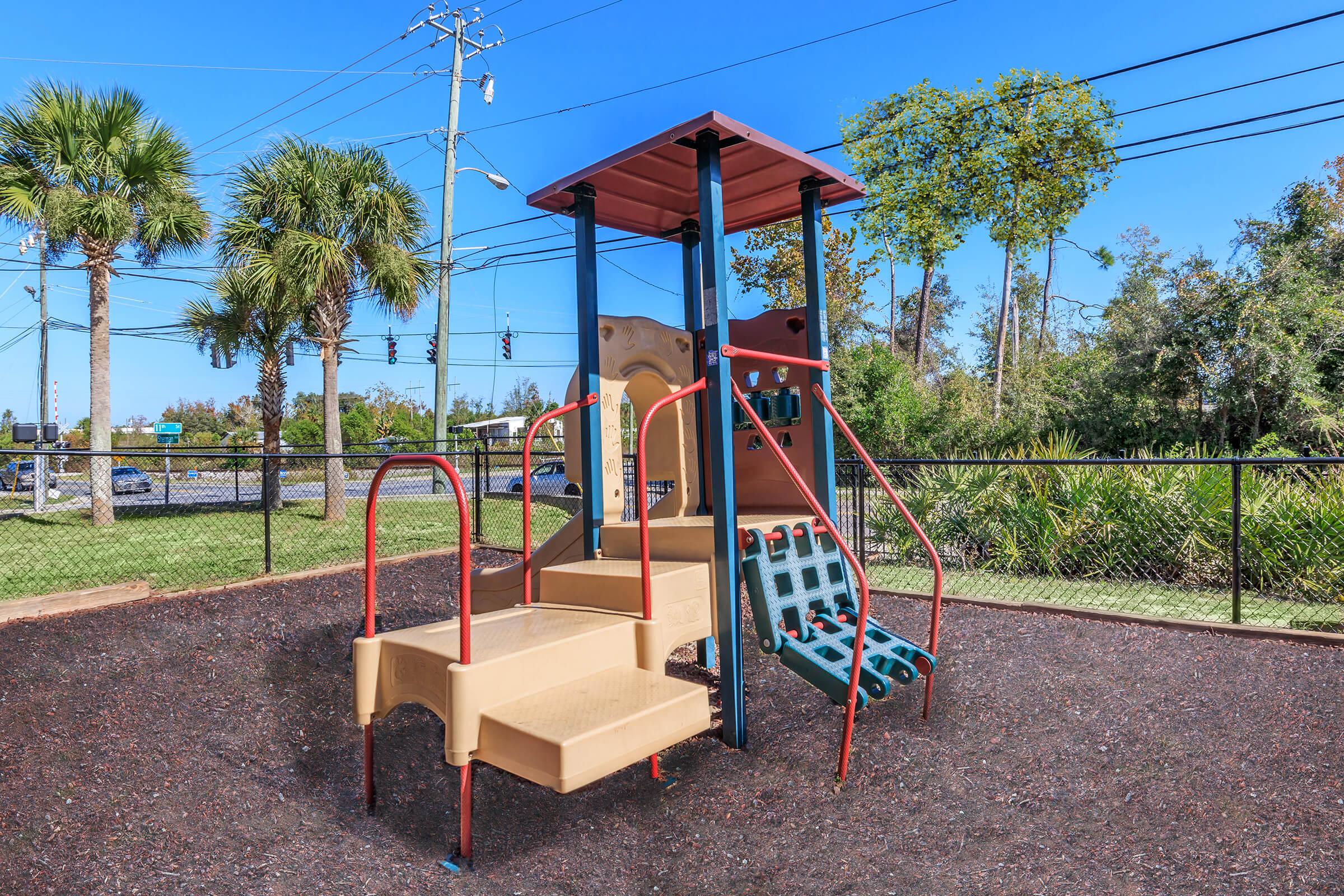 A colorful playground structure featuring a slide and climbing wall, surrounded by safety matting. Lush green palm trees and shrubs are in the background, with a clear blue sky above. In the distance, traffic lights and roads are visible beyond a fence.