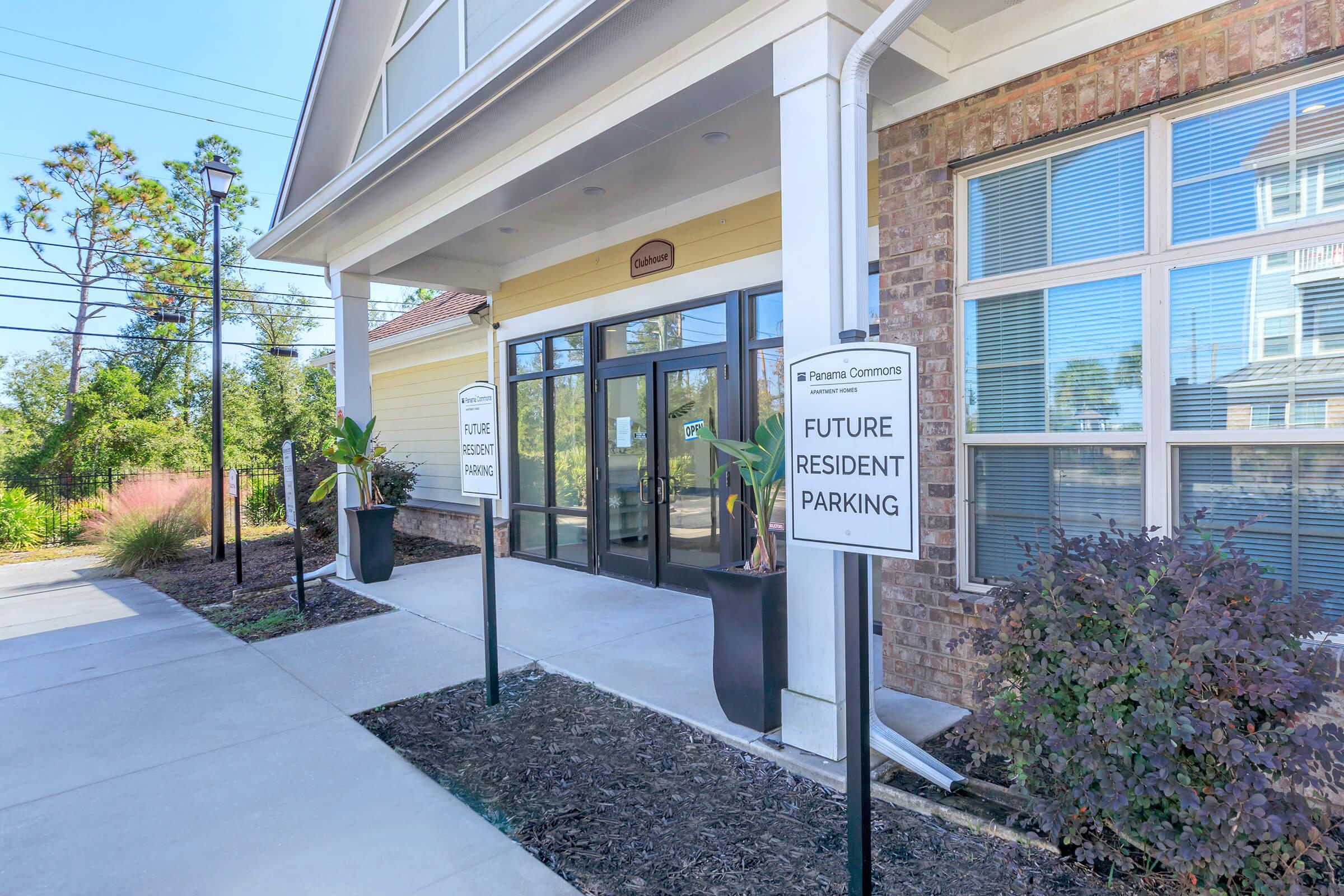 Exterior view of an apartment building entrance with large glass doors. Signs indicate “Future Resident Parking” flanking the entrance. The area is landscaped with plants and shrubs, and there are large windows showcasing a well-lit interior.