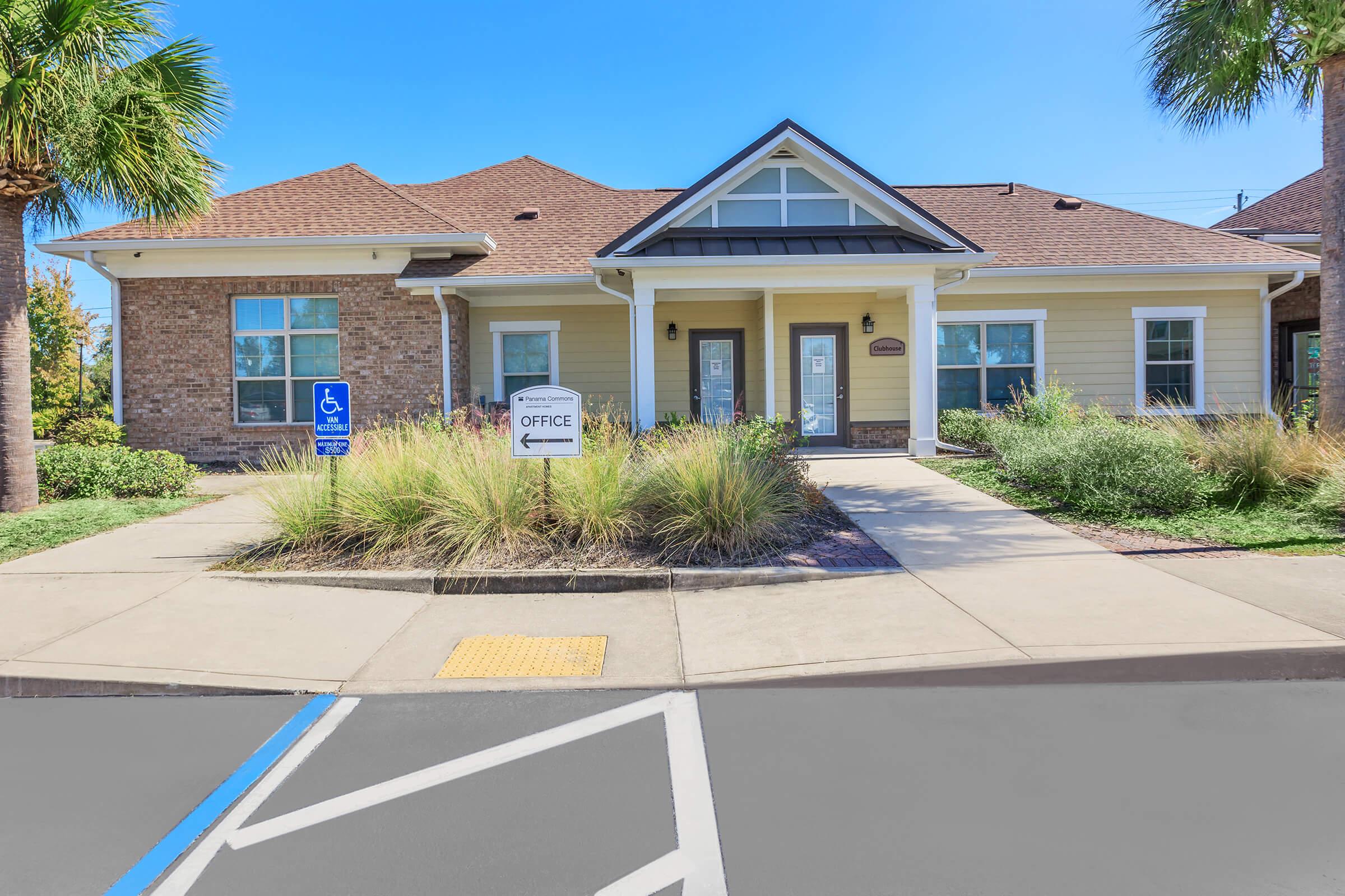 A one-story building with a combination of brick and siding. It features a sloped roof, multiple windows, and a sign indicating "Office." There are palm trees and landscaped plants in front, with a blue disabled parking space visible to the left. Clear sky overhead.