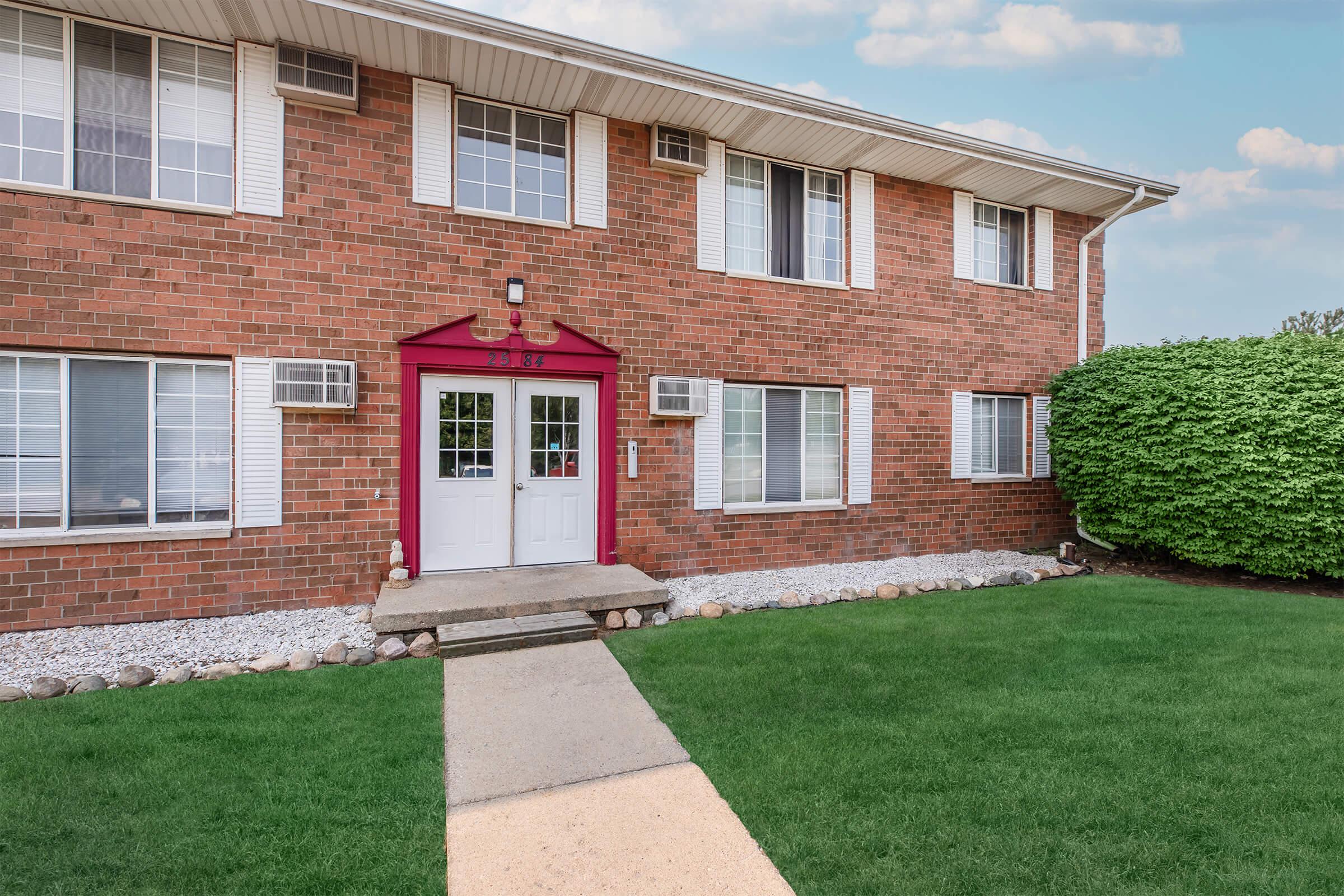 Front view of a brick apartment building with a well-maintained lawn. The entrance features a red canopy above double white doors, flanked by two air conditioning units. Windows are arranged symmetrically on either side, and there's a stone pathway leading to the door. Bushes are visible in the foreground.